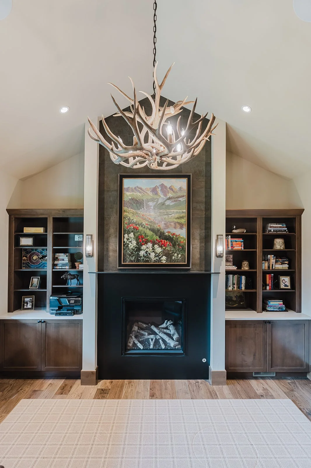 Living room with chandelier made of antlers, fireplace, landscape painting above fireplace, built-in bookshelves, and hardwood flooring.