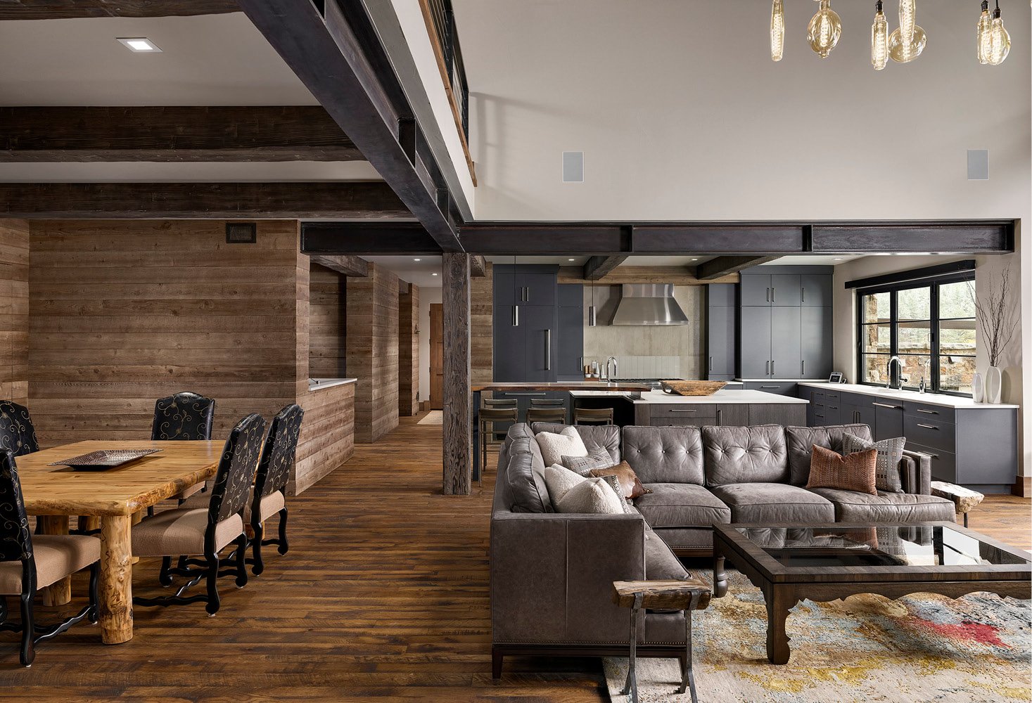 Open-concept living space with a rustic wooden dining table, black upholstered chairs, a gray sectional sofa with cushions, and a modern kitchen with gray cabinets, stainless steel range hood, and large windows for natural light.