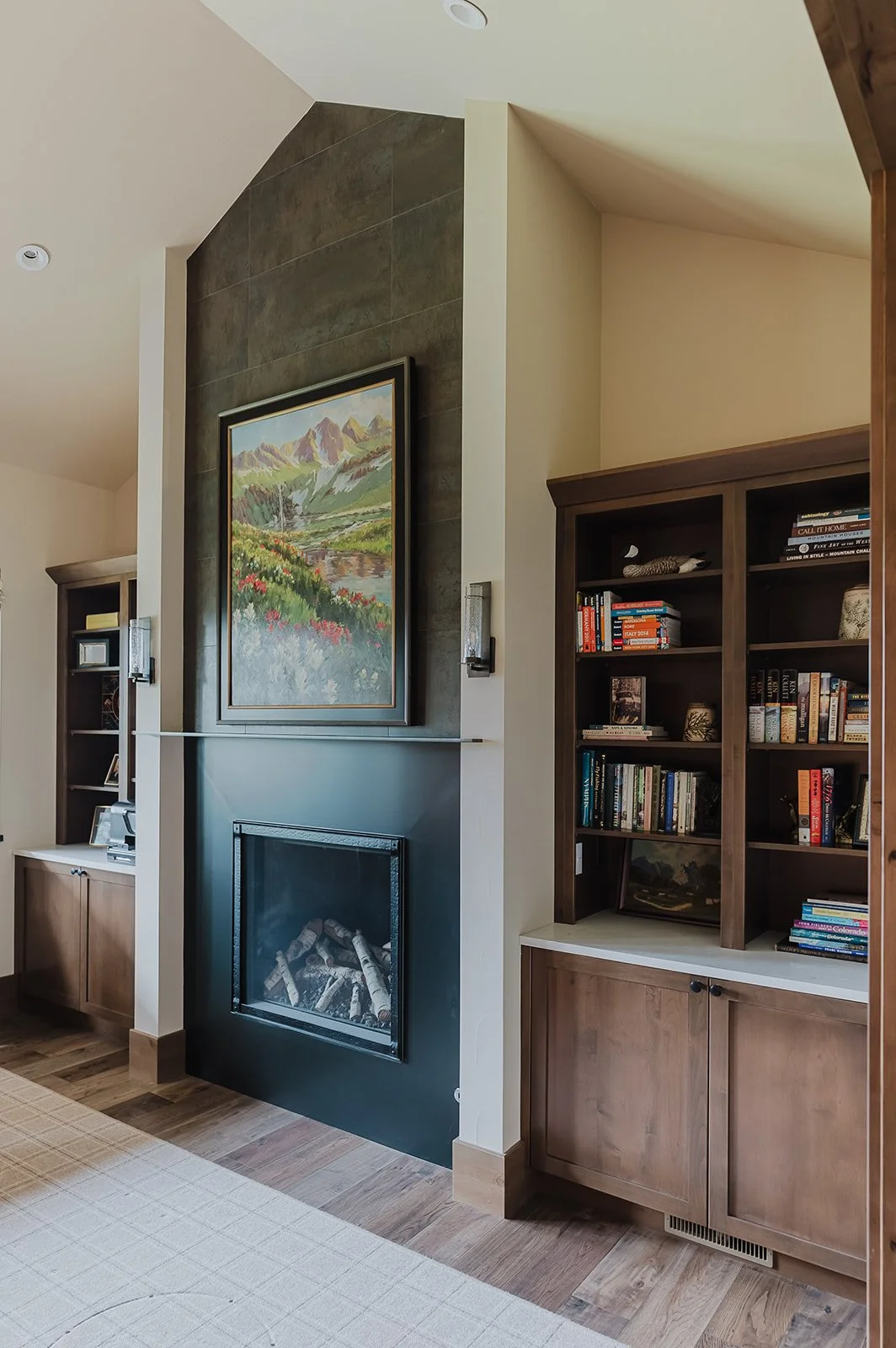 Living room corner with a black fireplace, a landscape painting above, bookshelves to the right, wood flooring, and beige walls.
