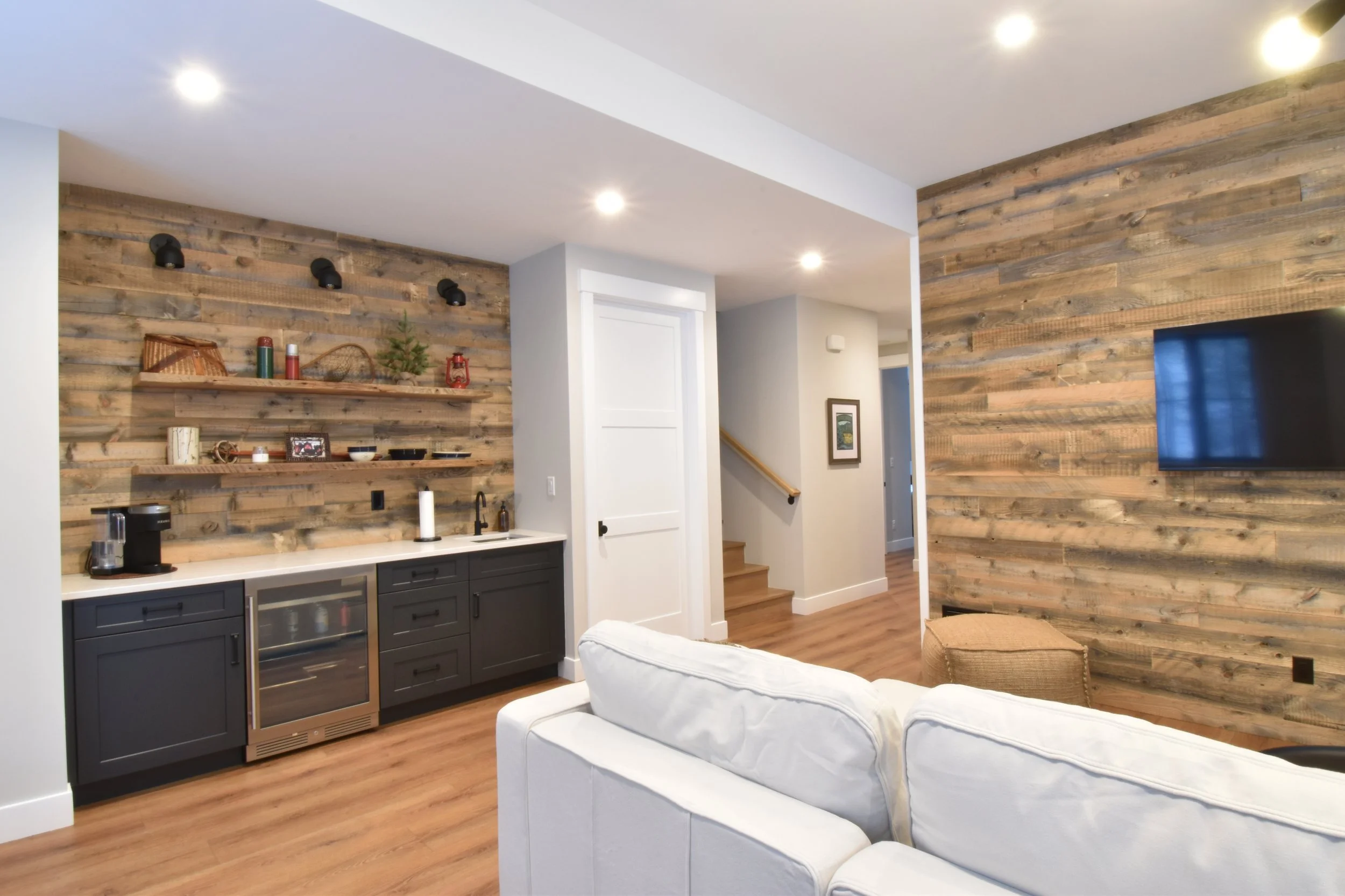Living area with a white sofa, wooden floors, and a wood-paneled accent wall. Open kitchen with dark gray cabinets, a wine fridge, and open shelves with decorative items. Recessed lighting and a flat-screen TV mounted on the wall.