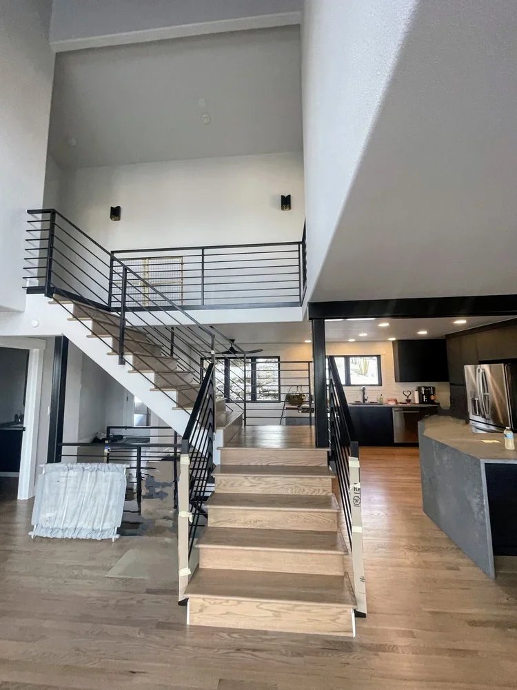 Interior of a modern home with a staircase, kitchen, and open living area with hardwood floors and metal railing.