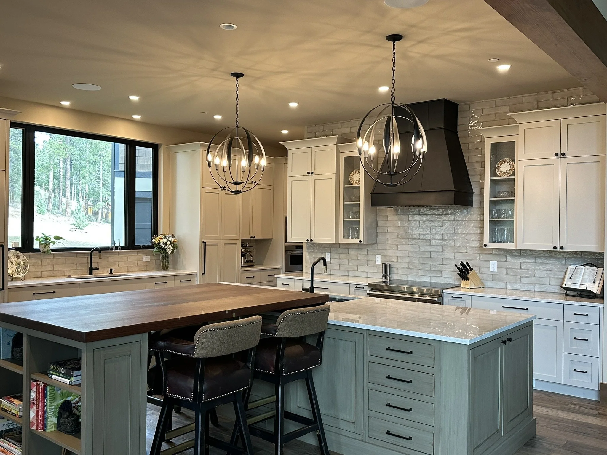 Modern kitchen with white cabinets, a large wooden island, black pendant chandeliers, a brick backsplash, a large window overlooking trees, and various kitchen utensils and decor.