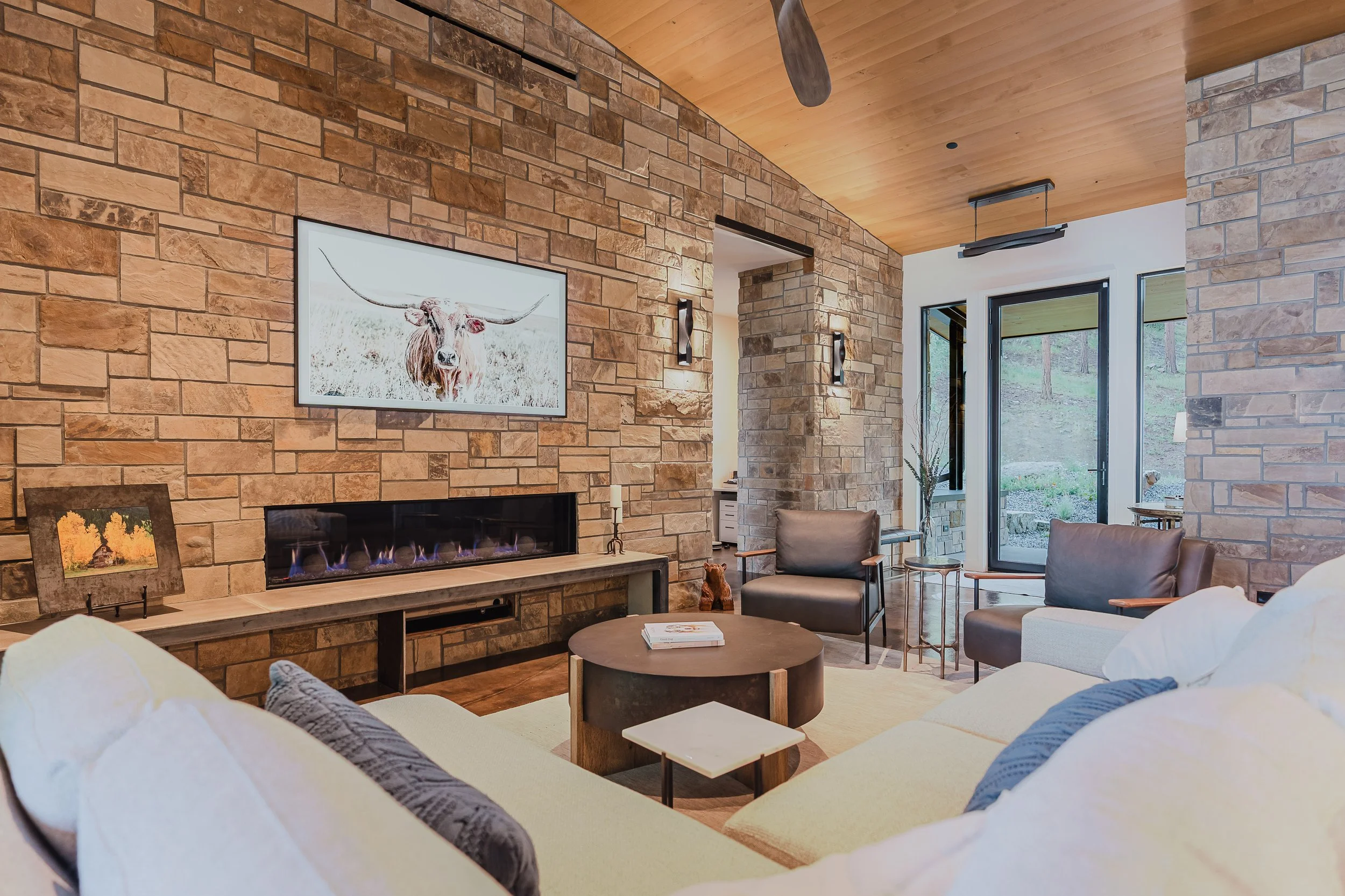 Living room with beige sofa, wooden coffee table, stone fireplace, TV, armchairs, artwork, and large windows showing a wooded yard.
