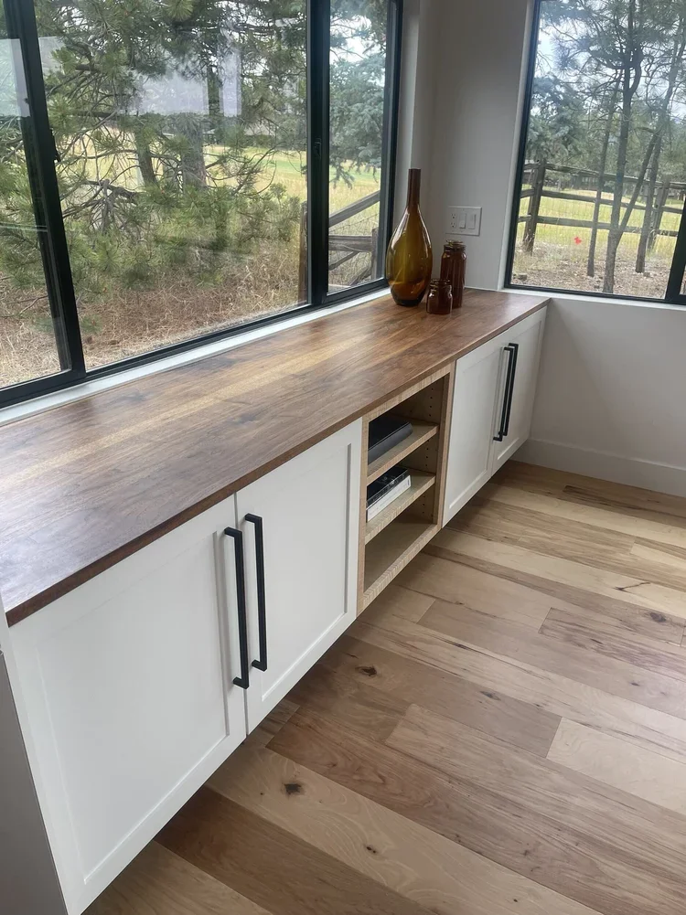 Wooden sideboard with white cabinet doors and open shelving, decorated with brown and amber glass vases, positioned in a room with large windows showing a wooded outdoor scene.