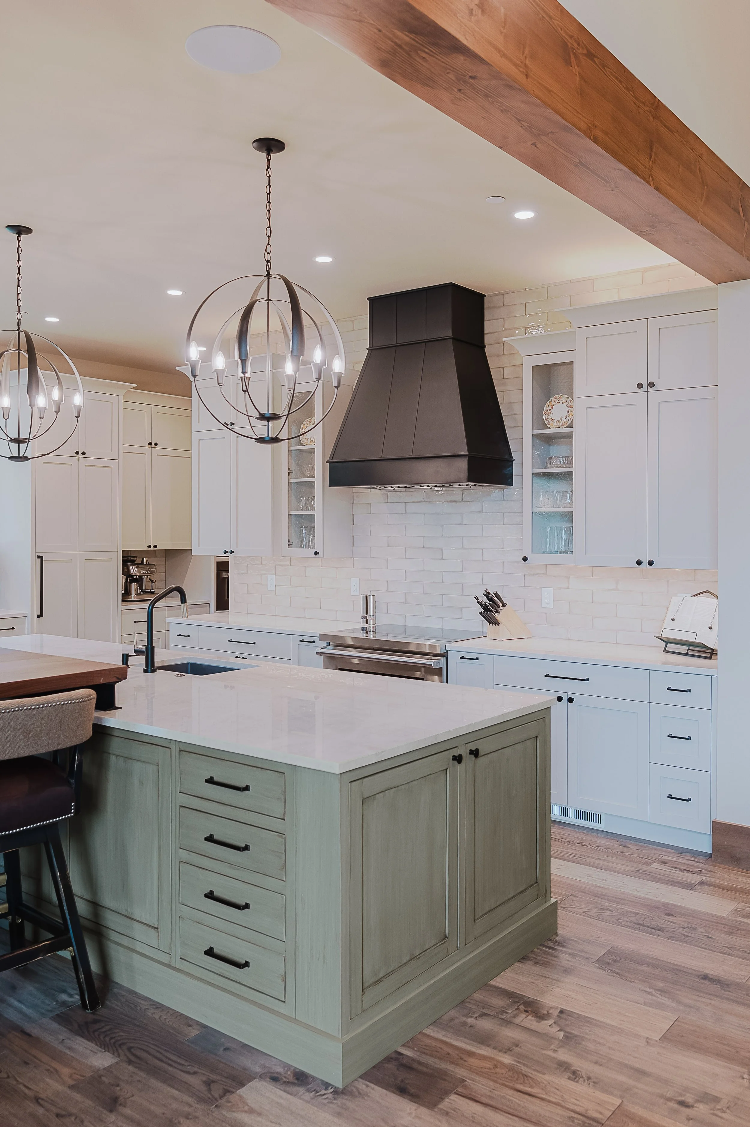 The image shows a modern kitchen with a large island painted in light green, featuring black handles and drawers. There are white cabinets with black knobs, a black range hood, and two spherical chandeliers hanging from the ceiling. The room has hardwood floors and a wood beam on the ceiling.