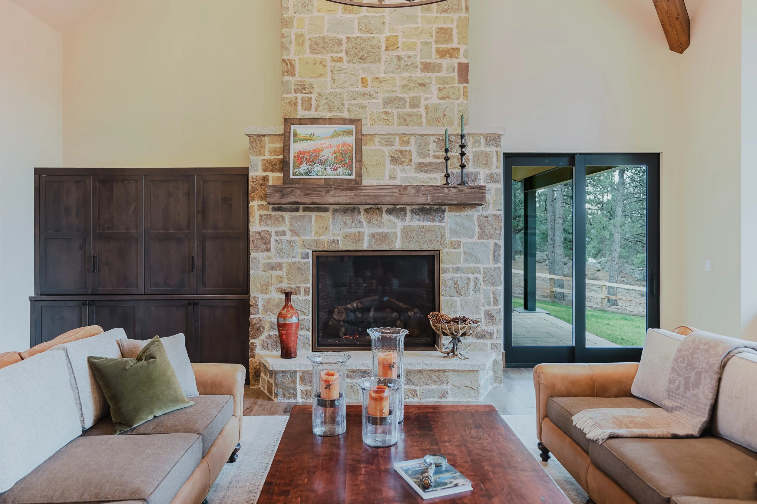 Living room with beige and brown couches, a stone fireplace with a wooden mantel, framed artwork, candles, and decorative items, large glass sliding door revealing a forest outside.