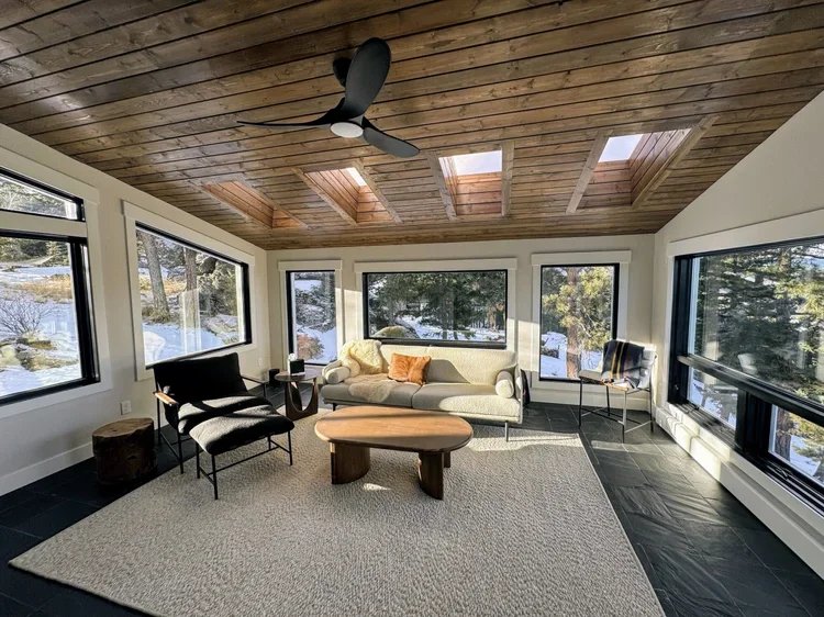 Living room with large windows showing snowy outdoor landscape, wooden ceiling with skylights, ceiling fan, white sofa with orange cushions, two black chairs, a small wooden coffee table, a side chair with striped cushion, and a beige rug on dark til
