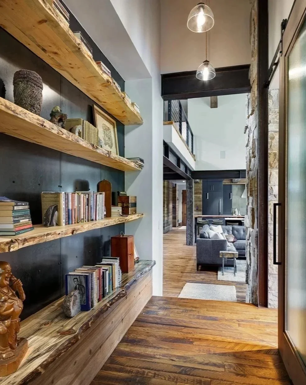 Interior view of a modern home with wooden flooring, open living space, gray sofa, bookshelves with books and decorative items, stone wall accents, and a staircase leading to a loft area.
