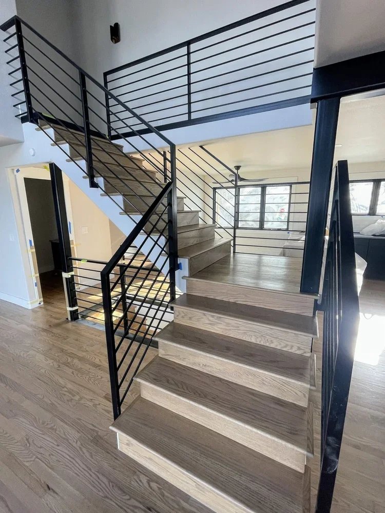 Interior view of a modern staircase with wooden steps and black metal railings in a well-lit home, with a second-floor landing and large windows in the background.