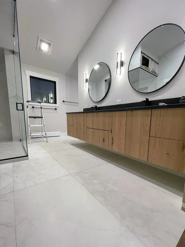 Modern bathroom with double oval mirrors above a black countertop, wooden vanity cabinets, a shower with glass door, window, small step ladder, and minimal decor.