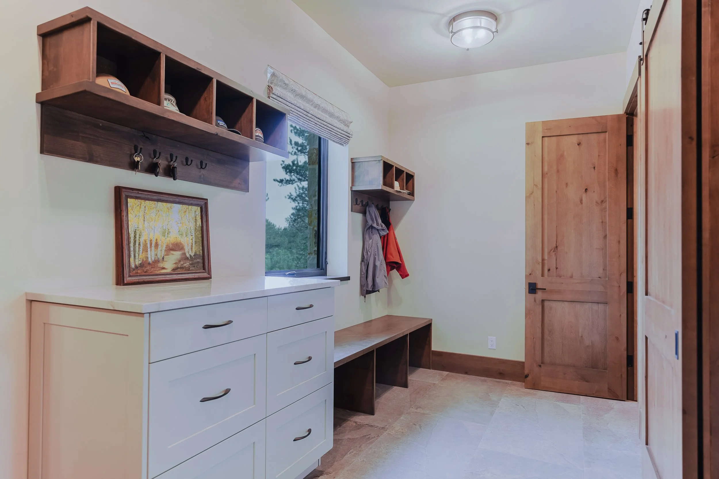 A modern entryway with a white sideboard, wooden hooks holding coats, and a wooden door, with a window showing trees outside.