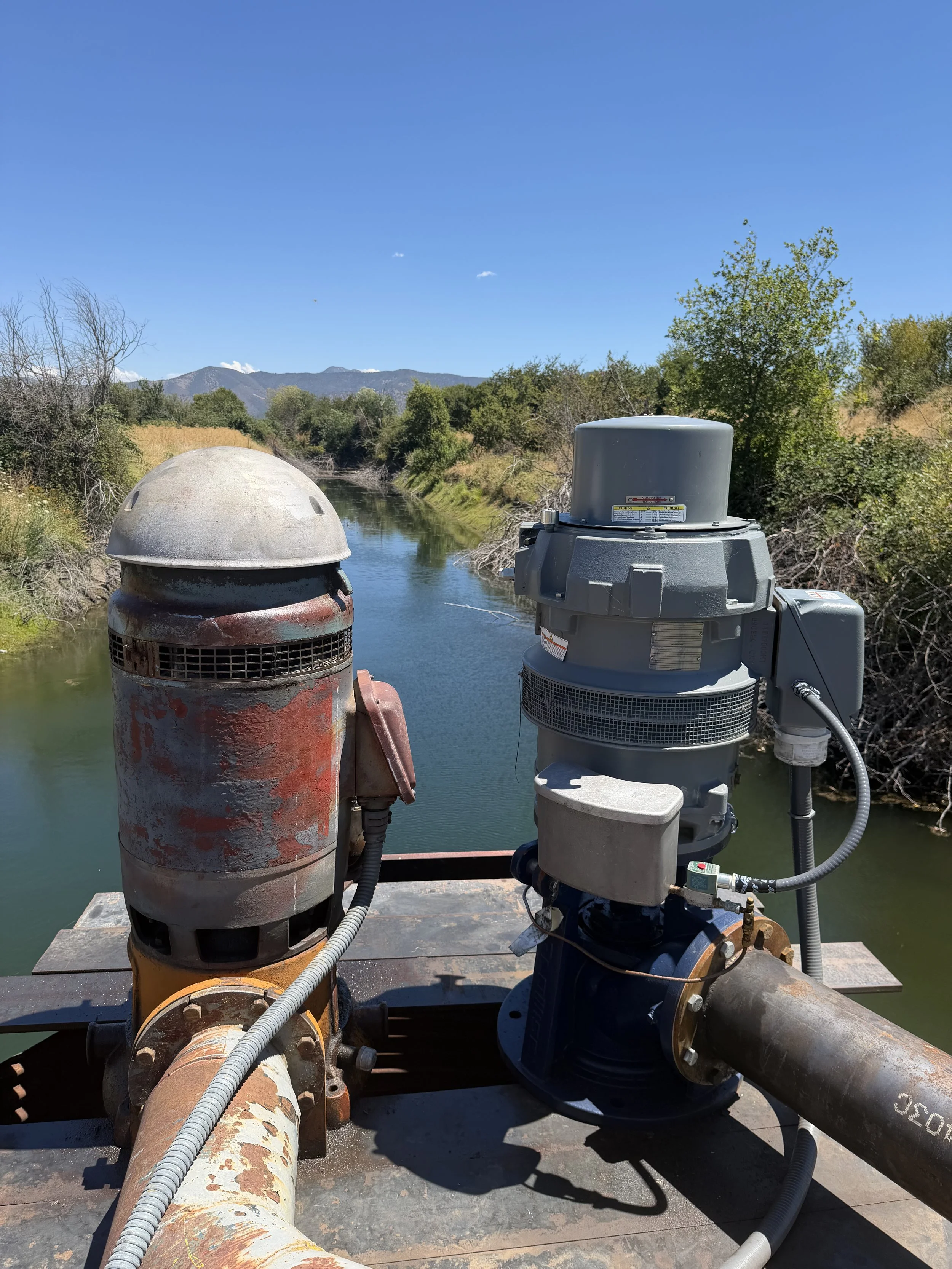 View from a boat showing industrial water pump system with pipes and machinery on a river, surrounded by trees and mountains under a clear blue sky.