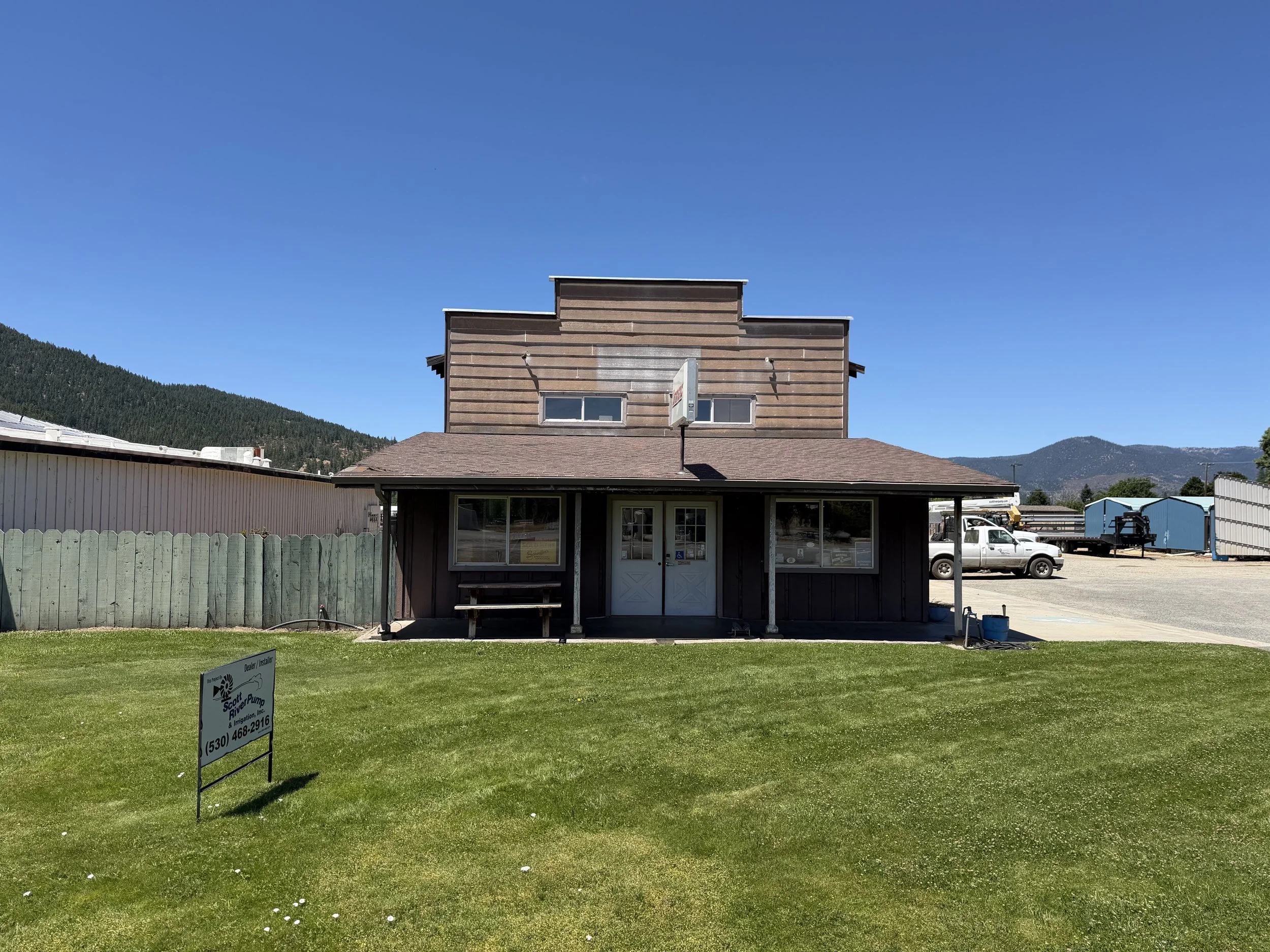 A two-story wooden building with a sloped brown roof, three windows on the upper floor, and a glass door on the lower level. The building is surrounded by a green lawn with a sign in front and a white picket fence. In the background, there are mountains and a bright blue sky.