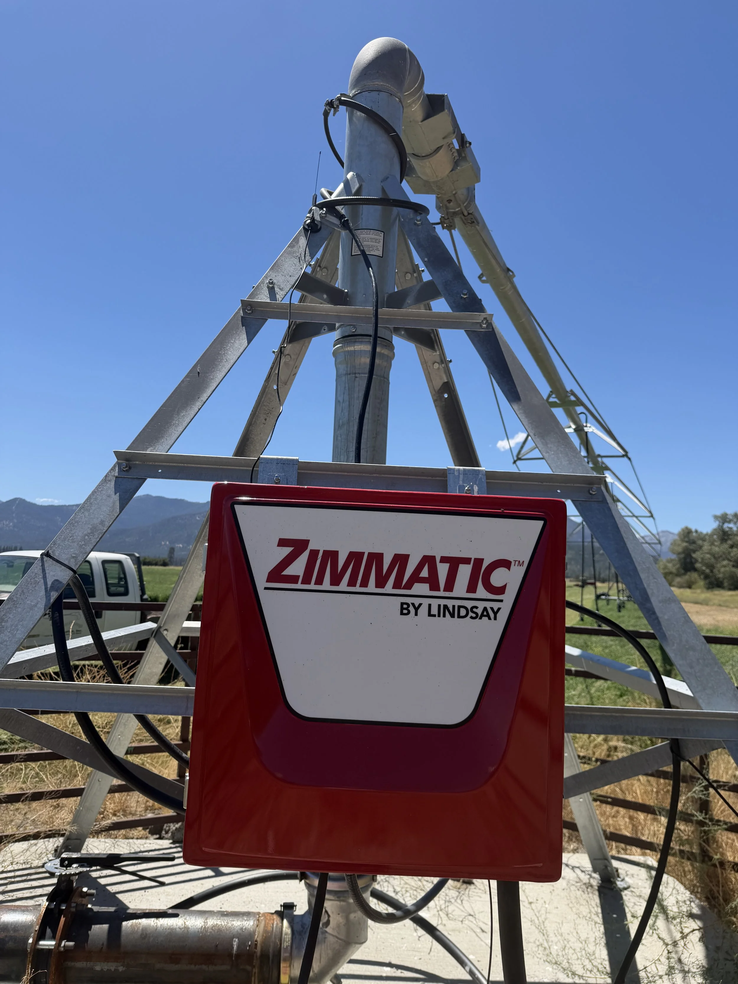 A tower with metal frame and machinery, with a red ZIMMATIC sign. The sky is clear and mountains are in the background.