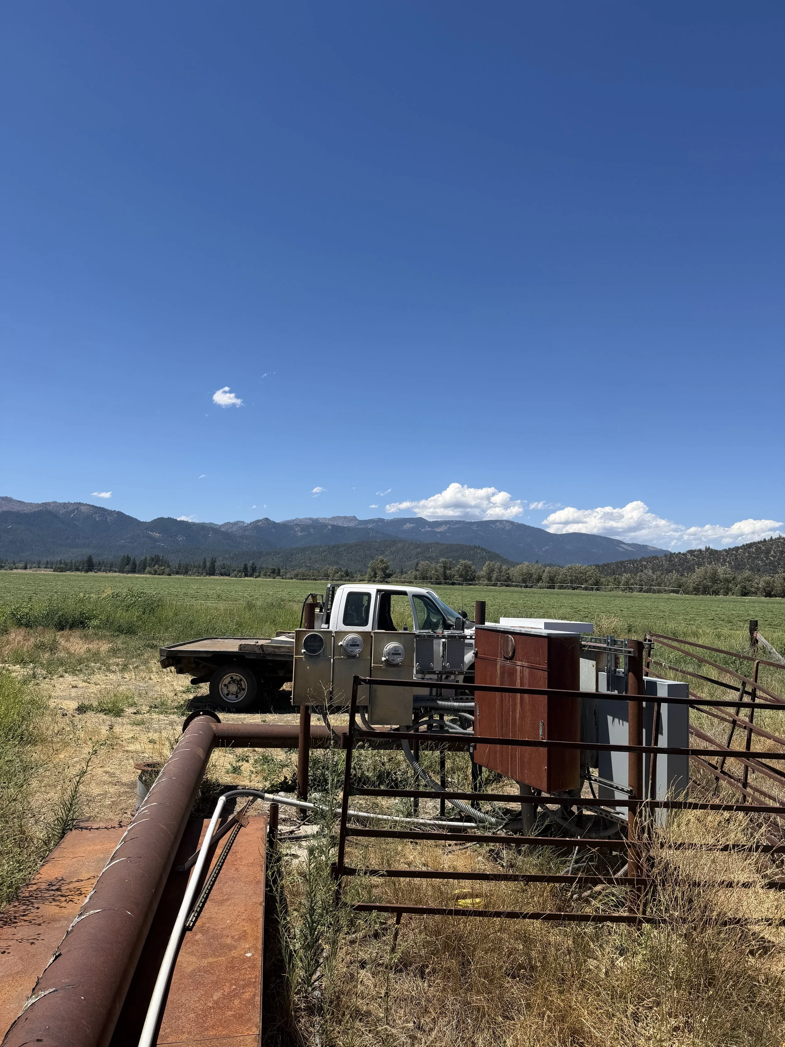 A white utility truck parked on a dirt area with industrial equipment, pipes, and wiring, overlooking green fields and mountains under a blue sky with scattered clouds.
