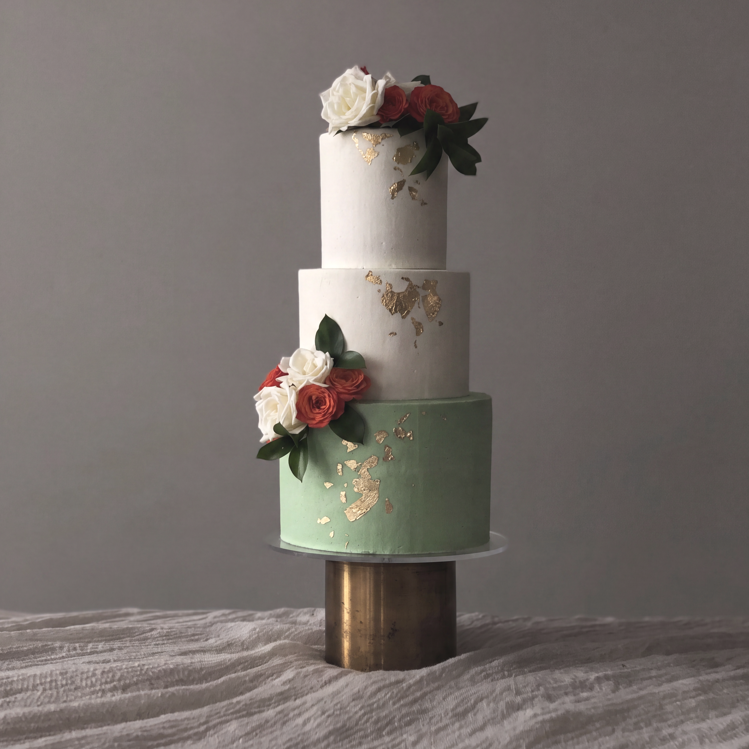 A two-tiered wedding cake decorated with roses, green leaves, and gold leaf accents.