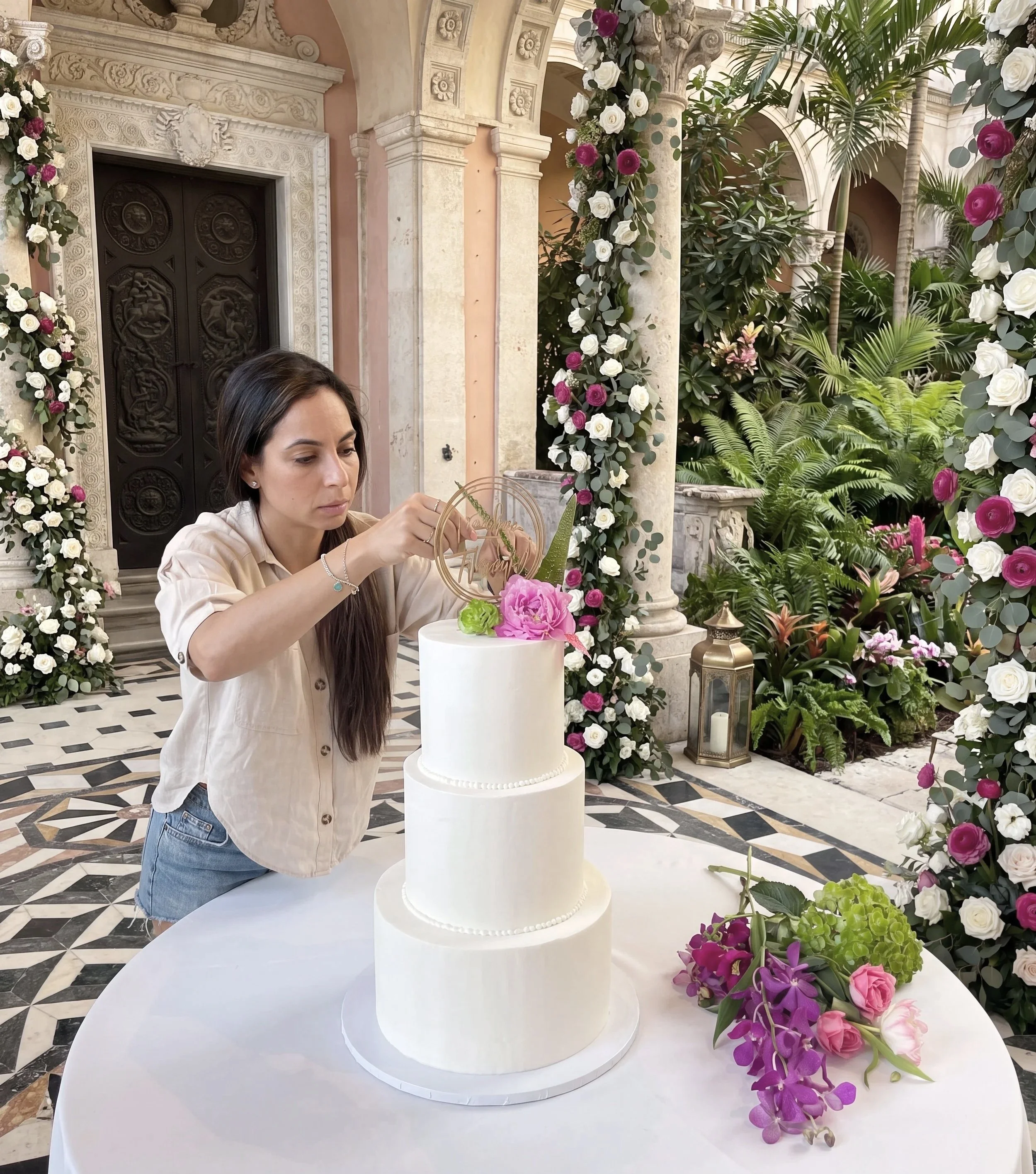 Chef Indira setting up and decorating a wedding cake with pink and purple flowers, surrounded by floral arrangements and lush greenery in an ornate hall in Coral Gables.