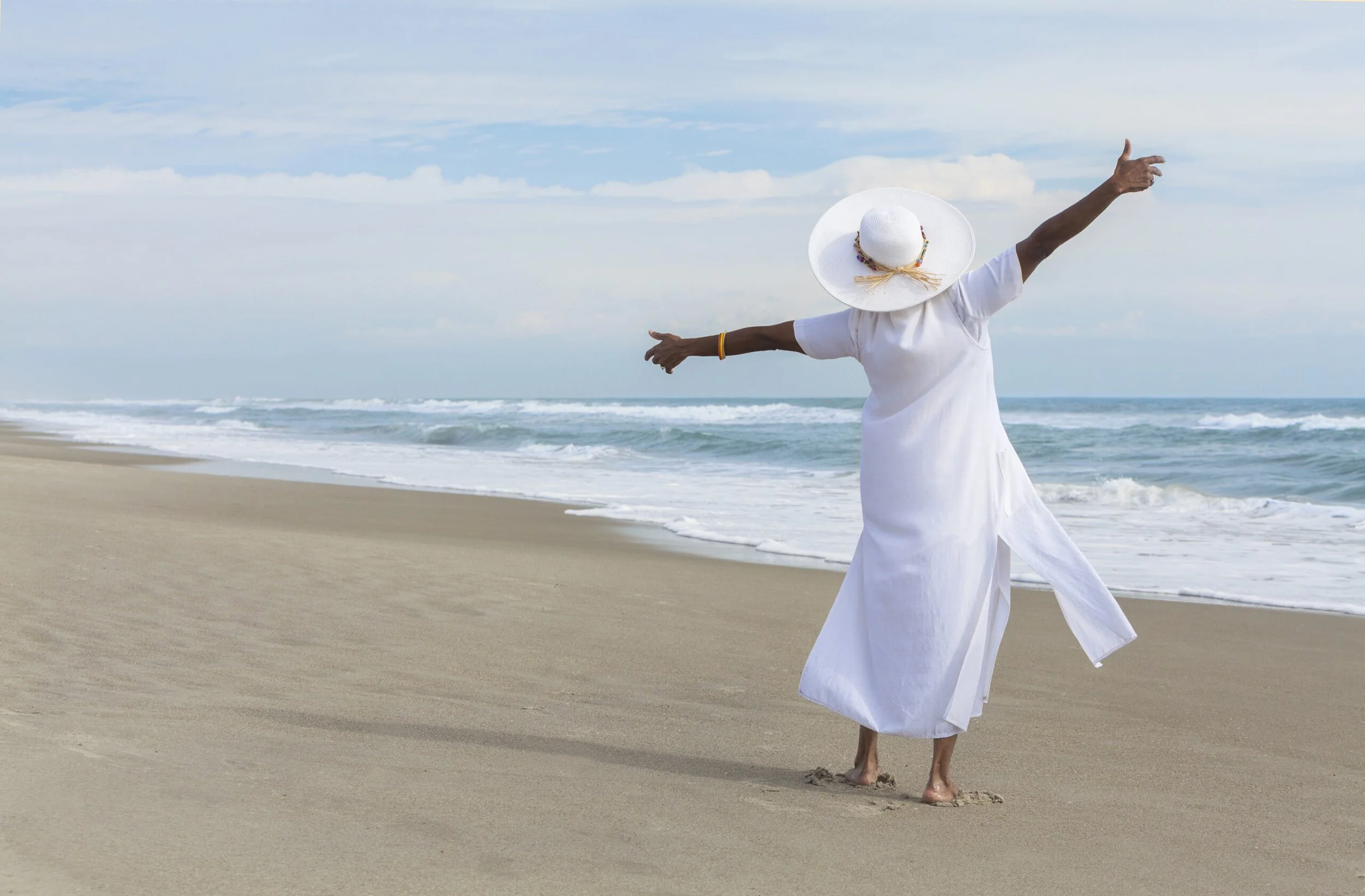 A black woman in a white dress and sun hat standing at the beach with arms wide open, embracing this next season of life