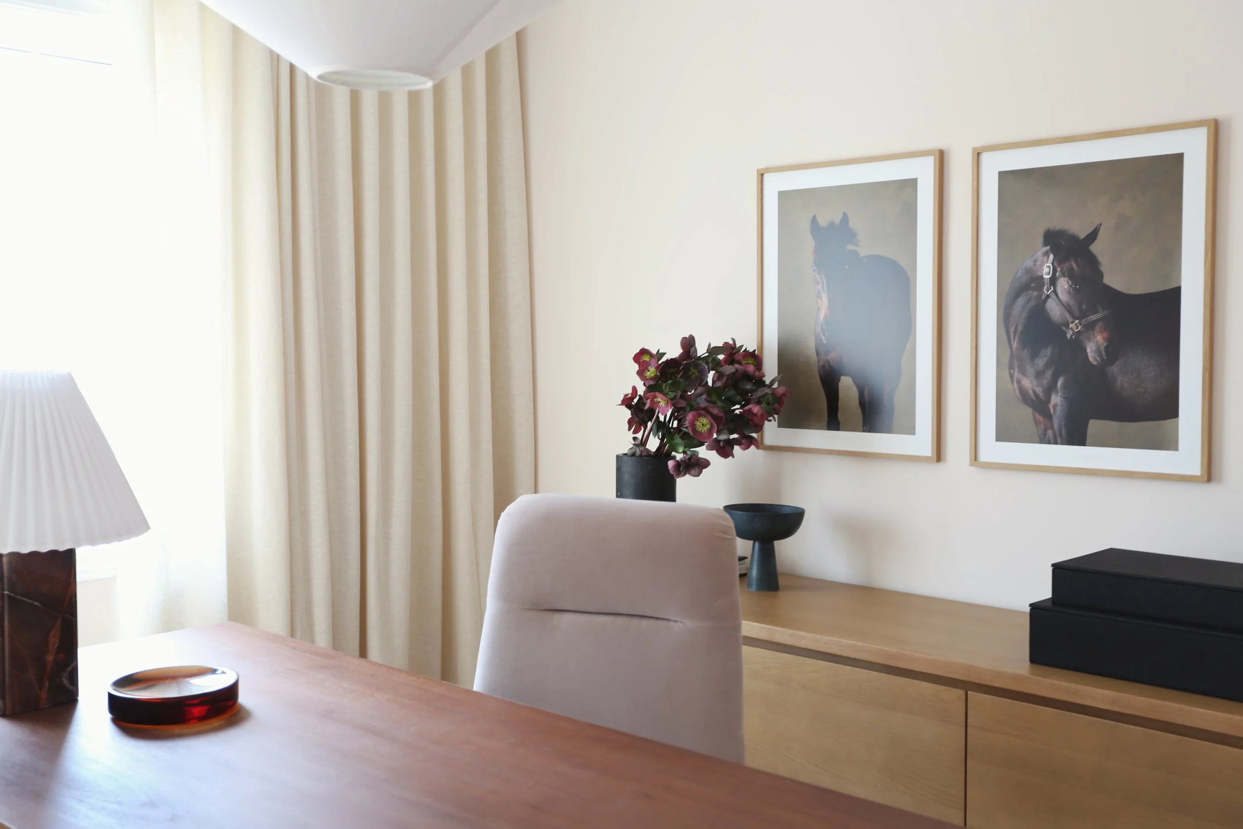 Living room corner with wall art featuring horses, beige curtains, a black vase with pink flowers, a table lamp, and a light pink chair.