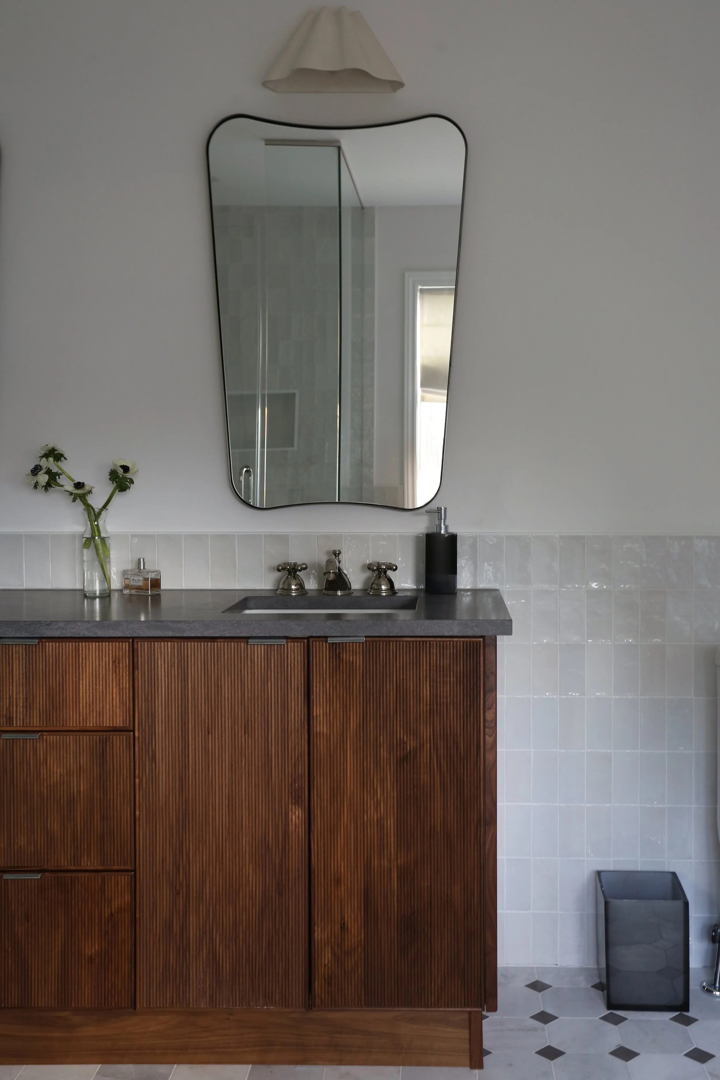 Bathroom vanity with a wooden cabinet, a gray countertop, a sink, and two faucets. On the left, there is a glass vase with white and black flowers. To the right, a black soap dispenser. In the background, a mirror above the vanity, a part of a shower, a window, a wall lamp, and a trash bin.