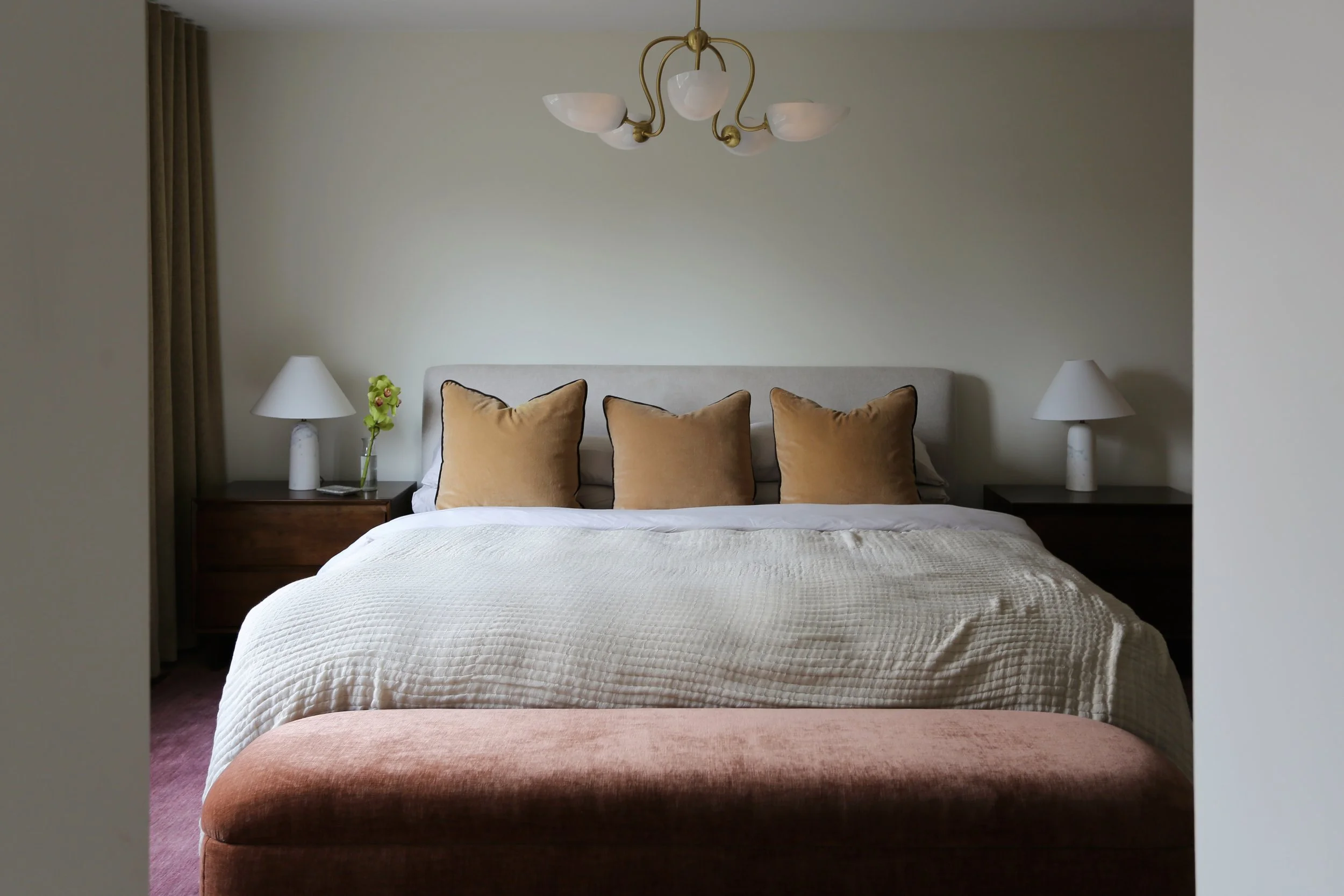 A bedroom with a bed topped with a beige quilt and four brown pillows. There are two matching nightstands with white lamps and a small flower vase on each side of the bed. A gold chandelier hangs from the ceiling, and beige curtains are partially visible on the left side.