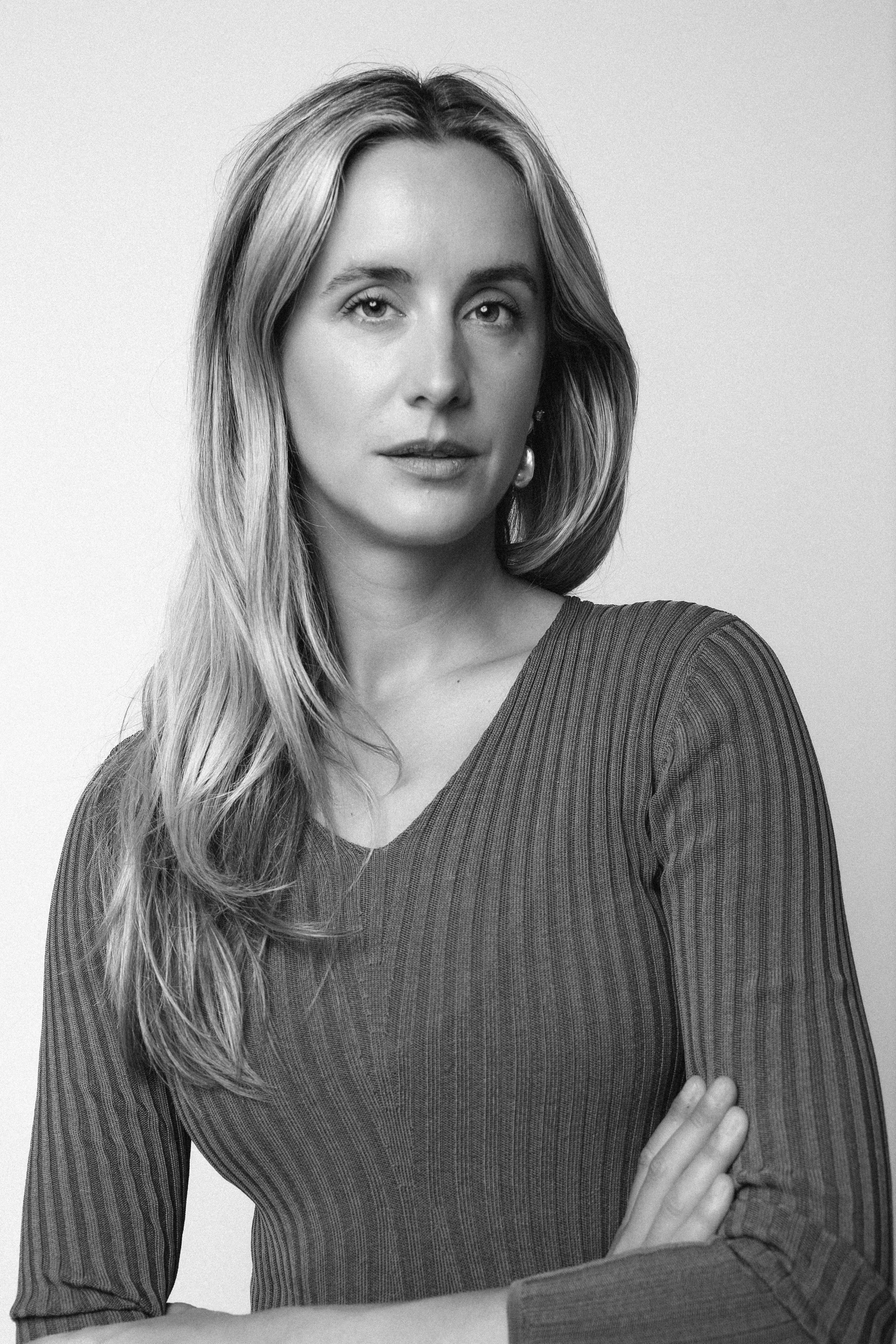 Black and white portrait of a woman with long, wavy hair, wearing a ribbed sweater, standing against a plain background.