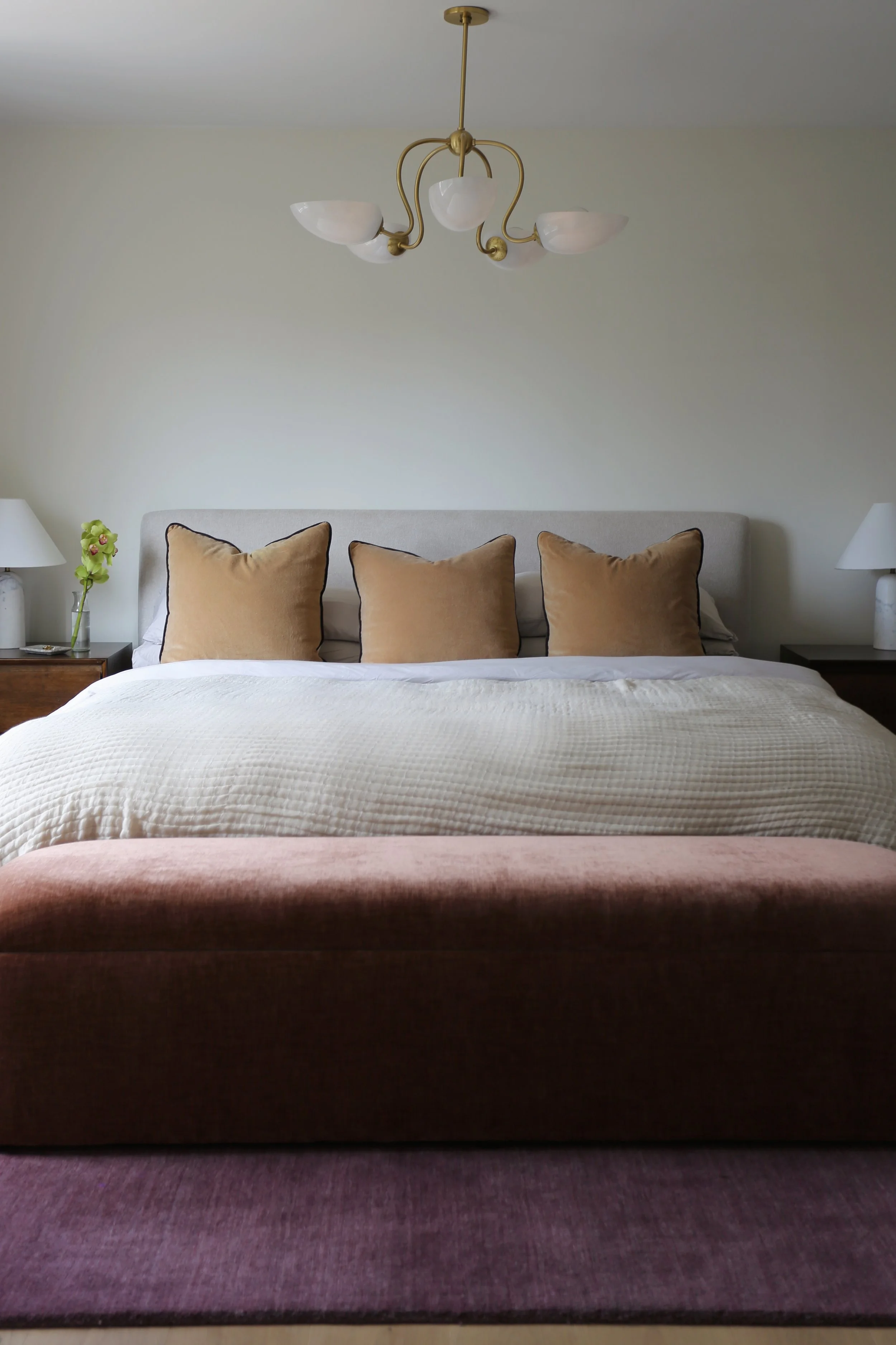 A bedroom with a large bed having a beige headboard and three beige pillows, a pink upholstered bench at the foot of the bed, white side tables with lamps, a tall lamp with a white shade and a green plant on the left side, and a ceiling light fixture with five curved brass arms and white glass shades.