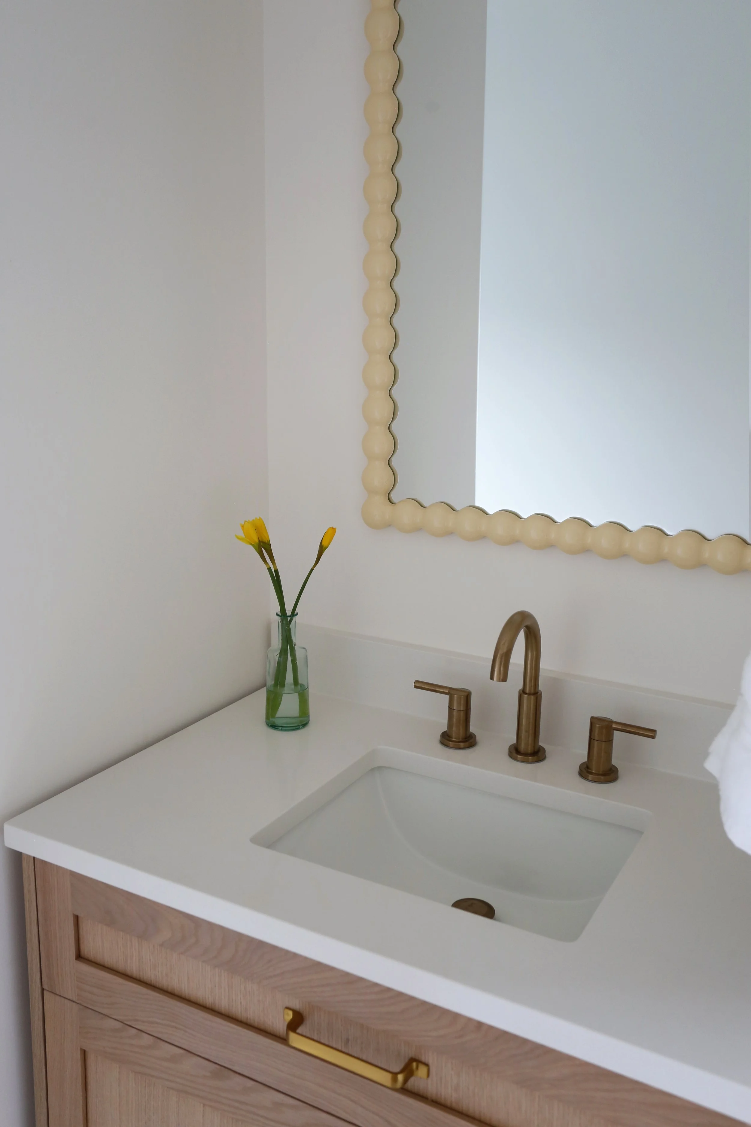 A bathroom vanity with a white countertop, a built-in sink, a wooden cabinet with a brass handle, a gold faucet, a mirror with a beaded frame, and a glass vase with yellow flowers.