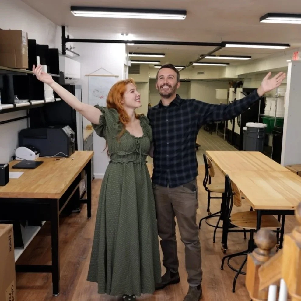 A man and woman standing in a room with their arms raised, smiling happily at each other, surrounded by tables and shelves in what appears to be a workplace or studio.