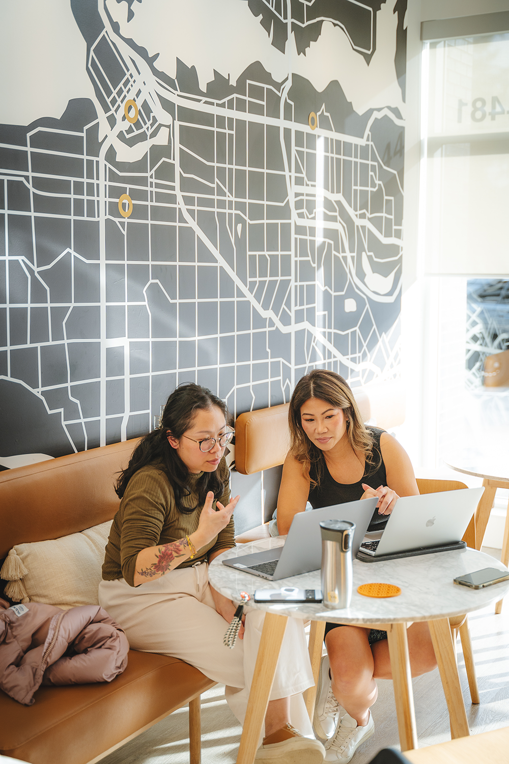 Two women sitting at a round marble table in a coffee shop working on laptops, with a large map mural on the wall behind them, and sunlight coming through a window.