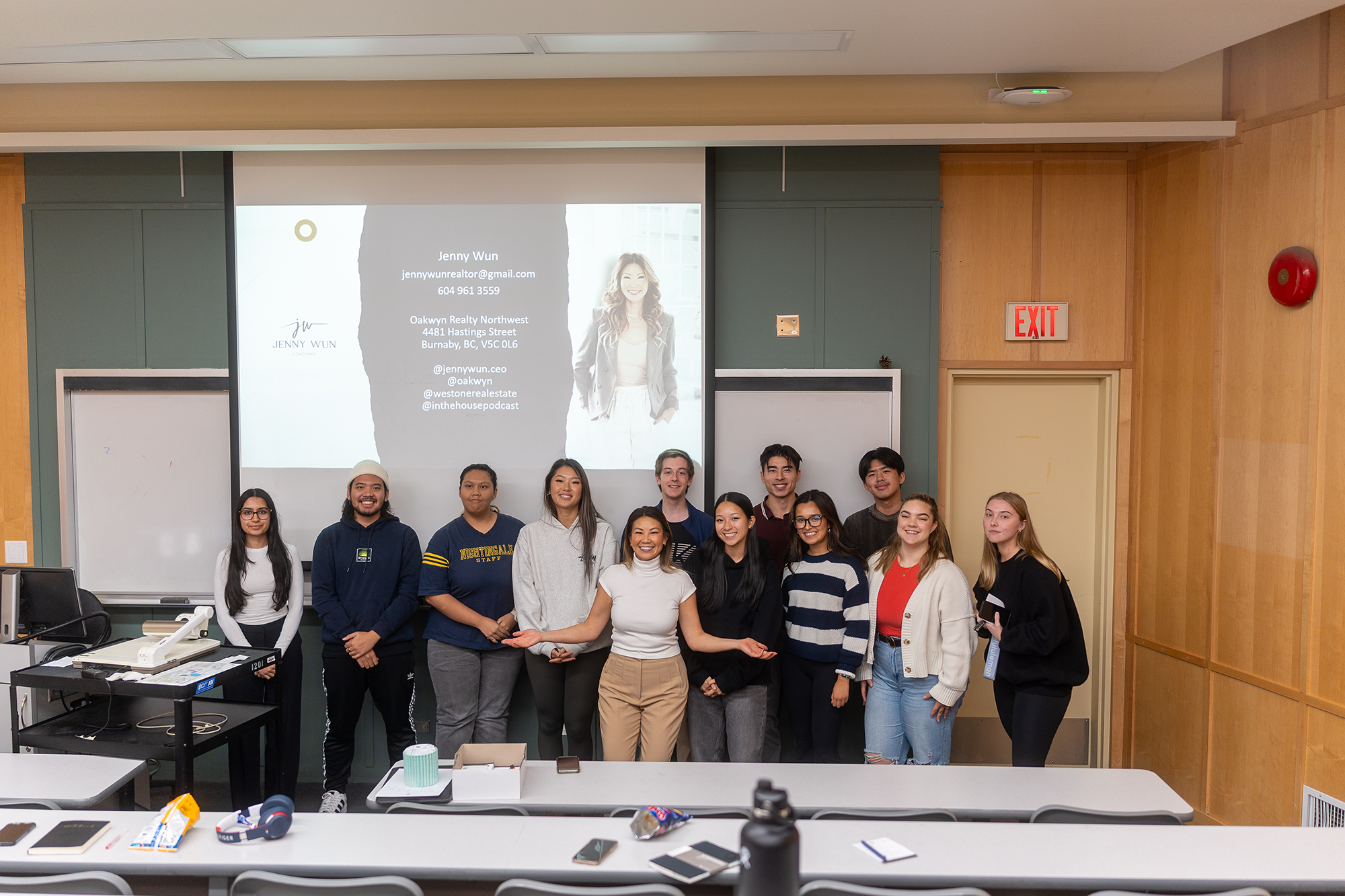 Group of diverse young people standing in a classroom in front of a projection screen displaying contact information for Jenny Wun, a realtor. The classroom has desks with various items including phones and notebooks, and a whiteboard is partially visible. The group is smiling and dressed casually.