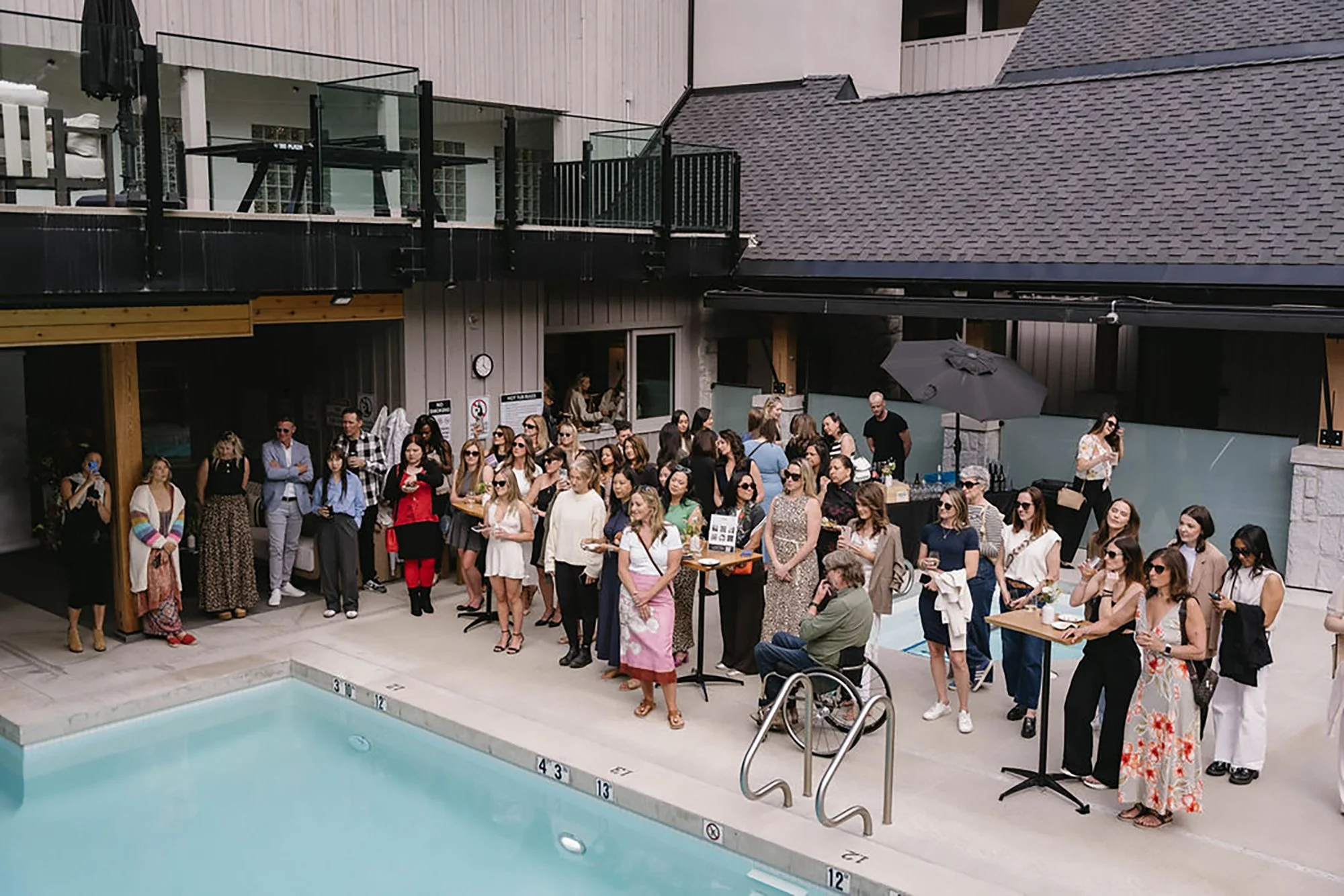 People gathering poolside at an outdoor event near a swimming pool with a crowd standing and sitting, some wearing sunglasses and holding drinks, in a modern venue with cabins and an outdoor patio area.