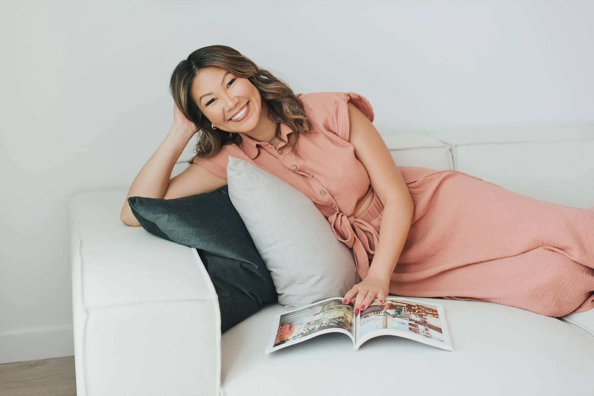 A woman with brown hair and a pink top relaxing on a white sofa, smiling and looking at a magazine.