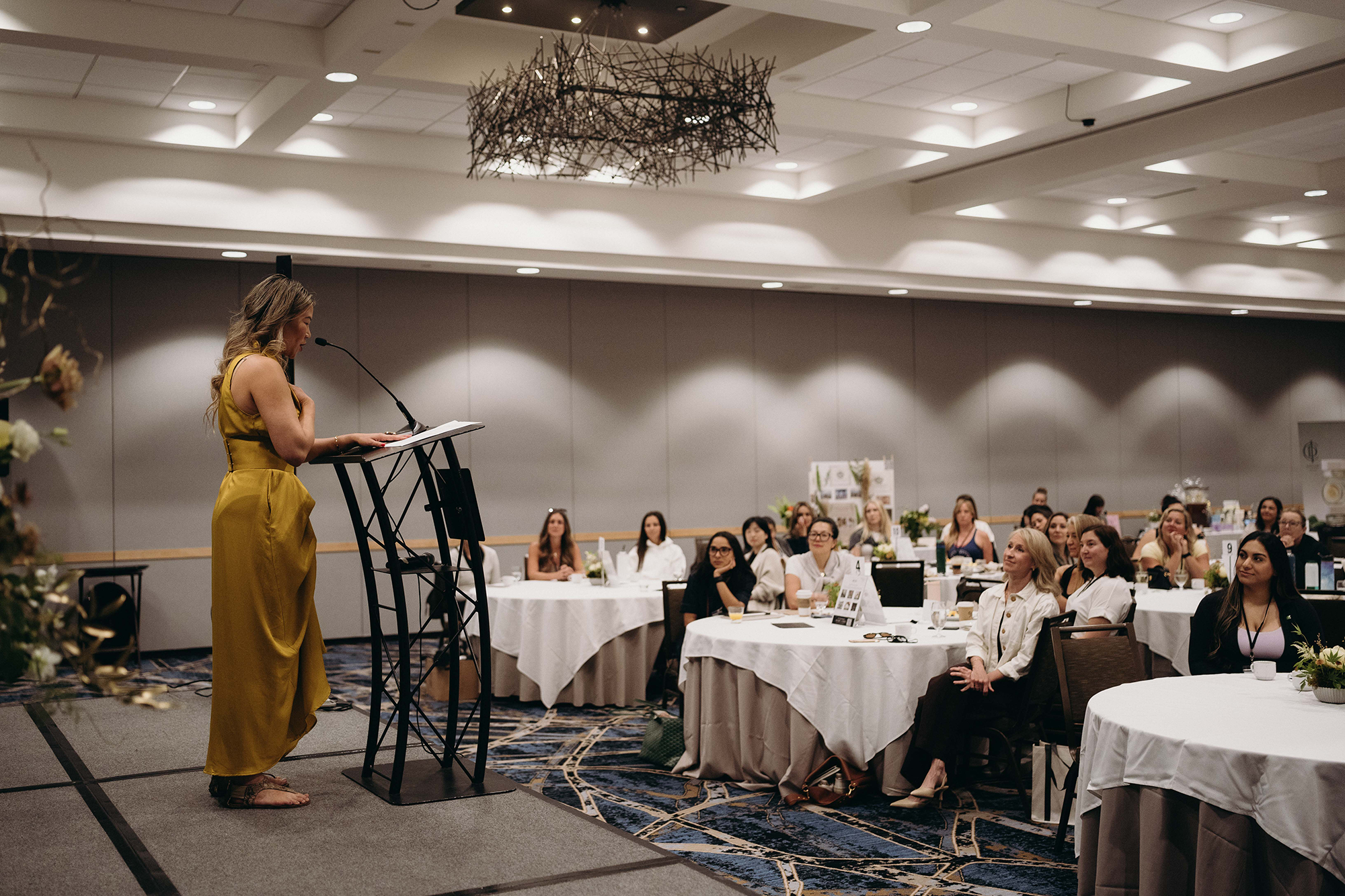 A woman in a yellow dress giving a speech at a podium in a large conference room with round tables and attendees seated, looking towards her.