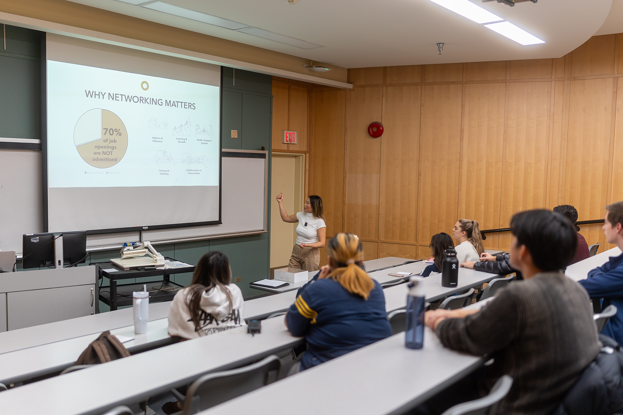 A classroom lecture with a female instructor standing in front of a projected slide that reads 'Why Networking Matters' with statistics and illustrations, and an audience of students attentively listening.