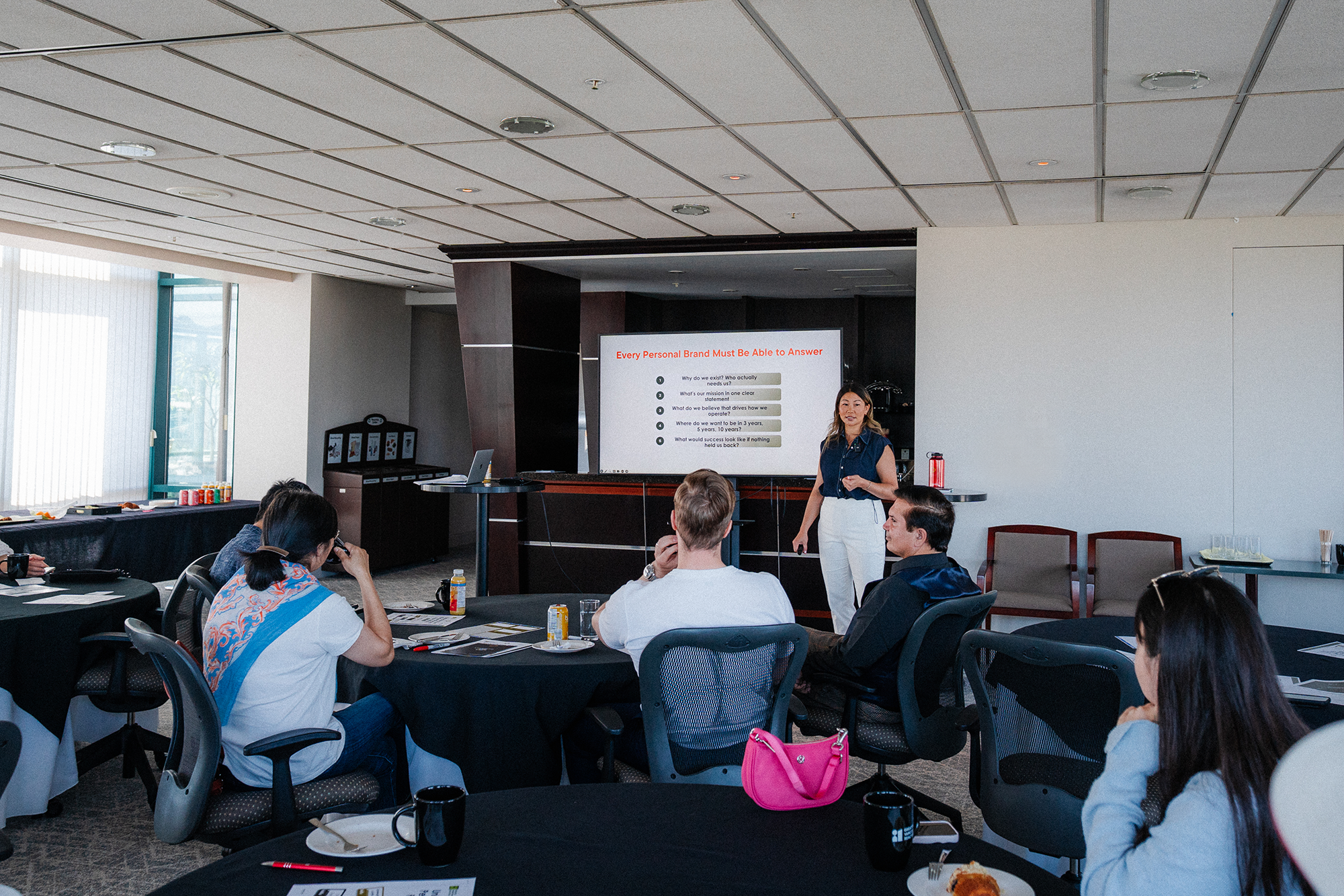 A woman presenting to a seated audience in a conference room, with a slide on the screen behind her titled 'Every Personal Brand Must Be Able to Answer'.