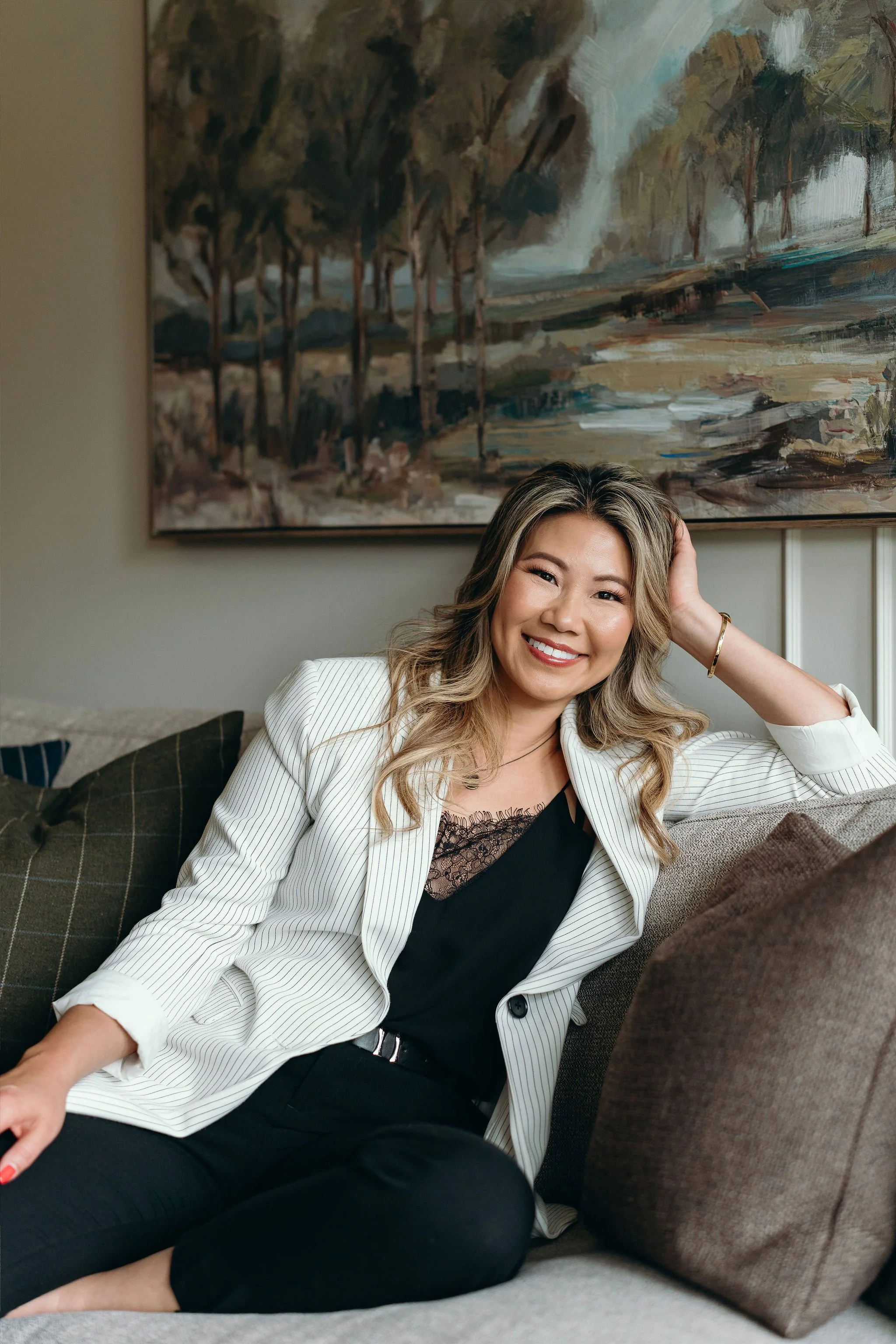 A woman sitting on a couch, smiling, with her hand resting on her head. She has wavy blonde hair and is wearing a white striped blazer over a black top with lace detail. Behind her is a large landscape painting of trees and a river.