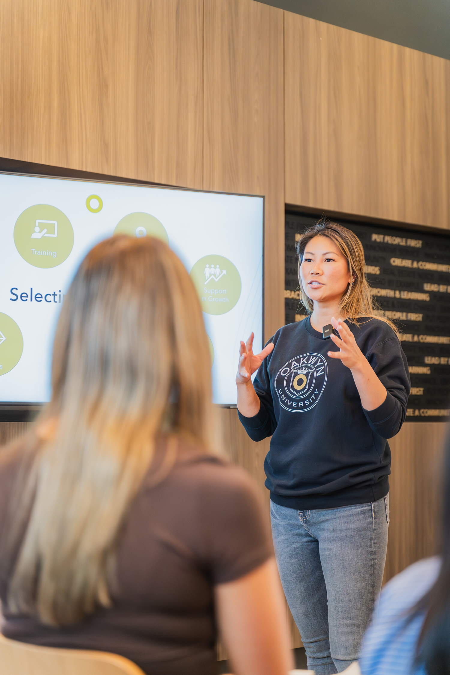 Woman giving a presentation to a group, standing next to a screen showing a diagram with the words "Training" and "Support in Growth" in a modern office setting.