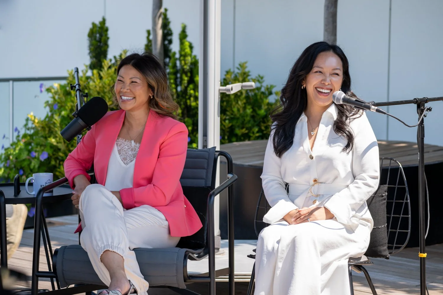 Two women sitting outdoors, smiling and laughing, with microphones in front of them. The woman on the left wears a pink blazer and white pants, and the woman on the right wears a white outfit. There are green plants behind them.