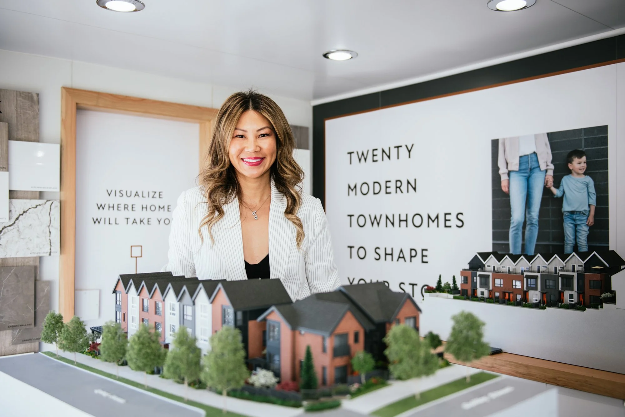 A woman with long wavy hair smiling in front of a model of a residential neighborhood, with posters and signs about modern townhomes in the background.