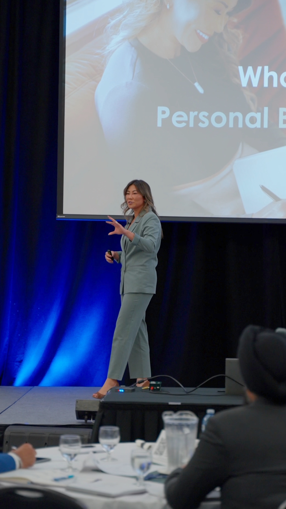 A woman in a gray suit giving a presentation on stage with a large screen behind her displaying a close-up of herself and the text "What Personal E." She is holding a clicker in her right hand, gesturing with her left, and standing in front of an audience at a conference or seminar.