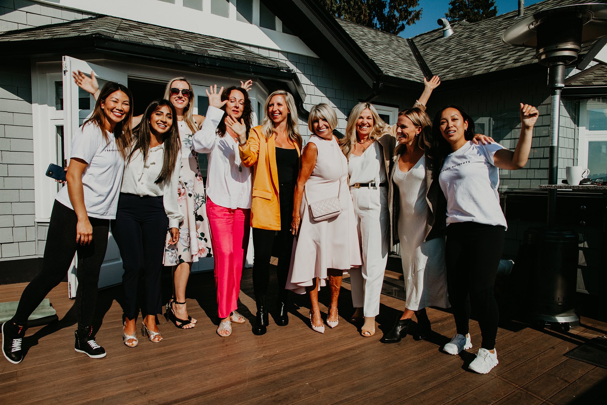 Group of women posing together outdoors on a wooden deck, smiling and making gestures, with a house and trees in the background.