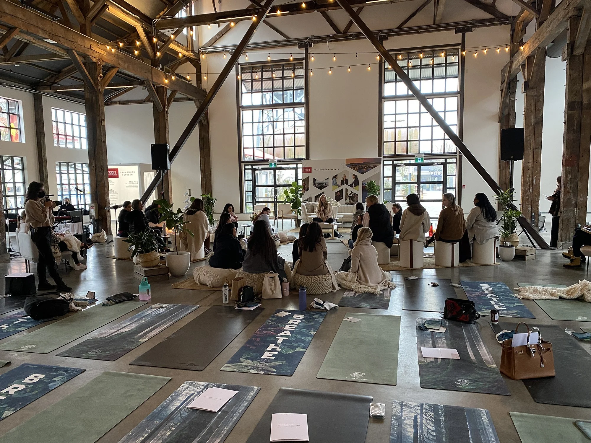 Inside a spacious, industrial-style event space with large windows and exposed wooden beams, where people are seated on cushions and chairs, preparing for a workshop or seminar, with yoga mats and printed materials laid out on the floor.