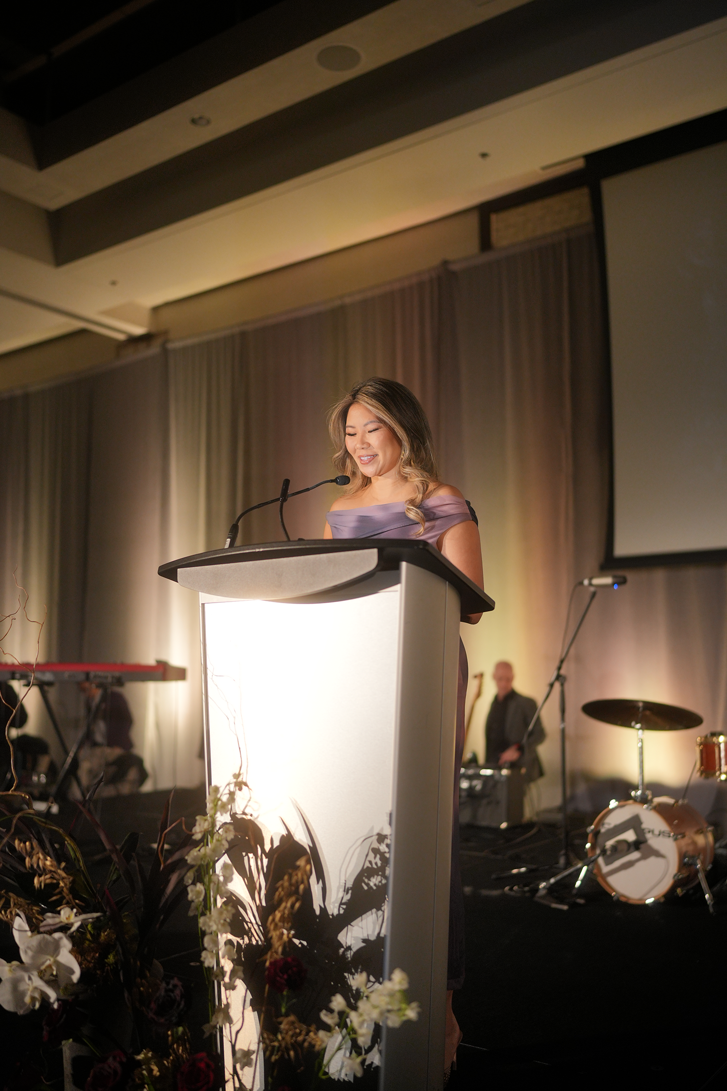 Woman standing at a podium, smiling and speaking into a microphone during a formal event, with musical instruments and a man in the background.