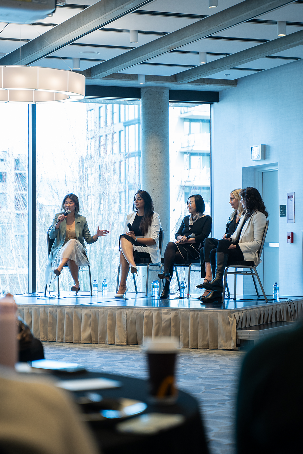 Five women seated on a panel stage in a conference room, with one woman speaking into a microphone. Large windows in the background show an urban building exterior.