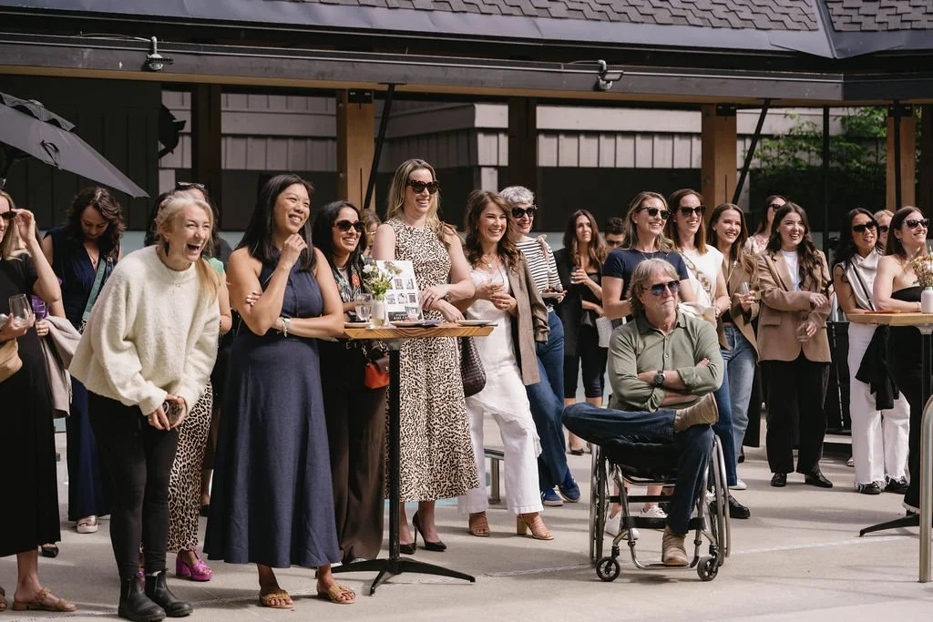 A group of women and one man in a wheelchair standing and laughing outdoors during a social event, with some holding drinks and others wearing sunglasses.