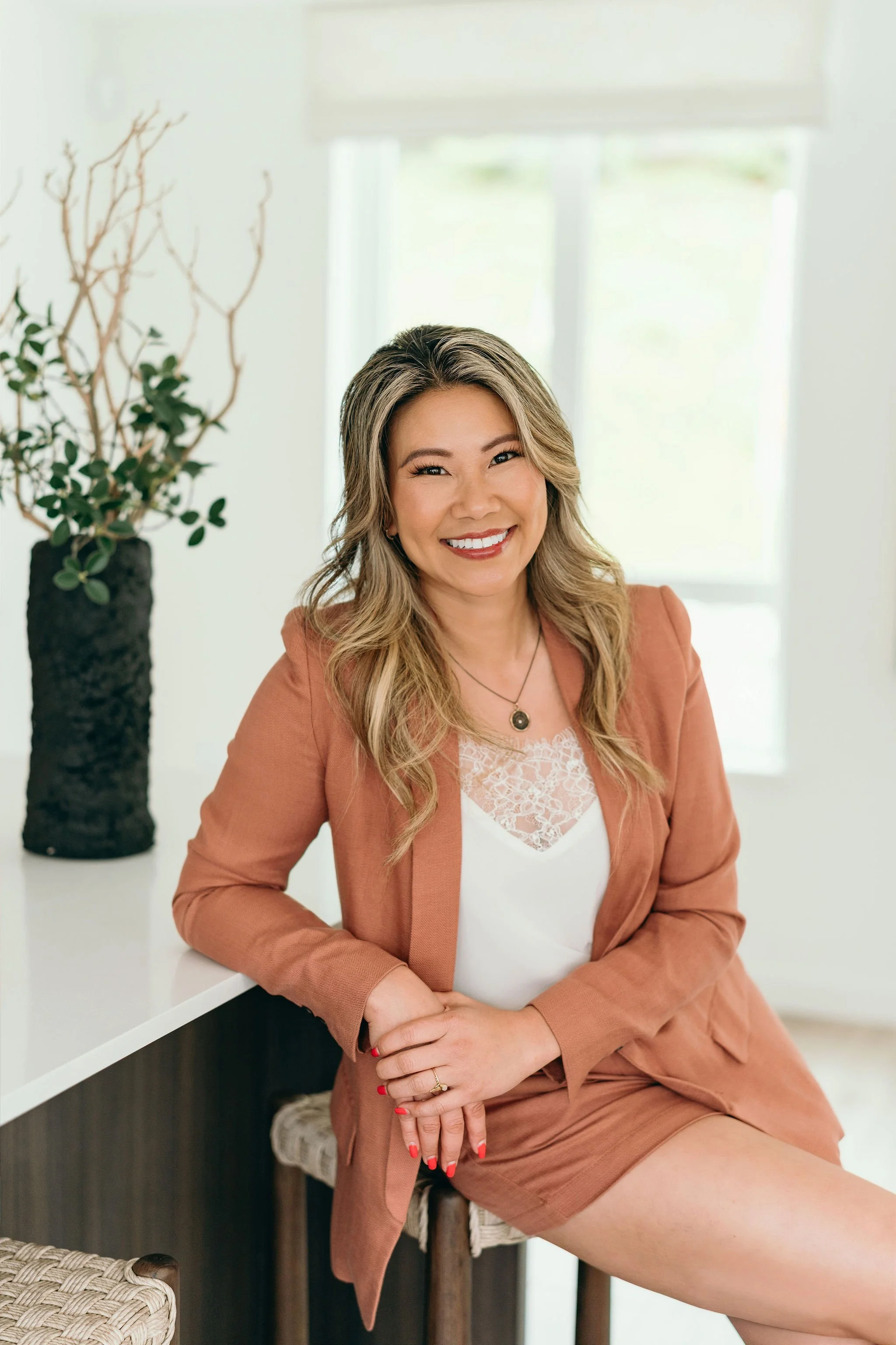 A woman with wavy blonde hair smiling, seated on a woven stool in a bright room, wearing a rust-colored blazer and matching shorts, with a white lace top and jewelry, with a tall plant and large window in the background.