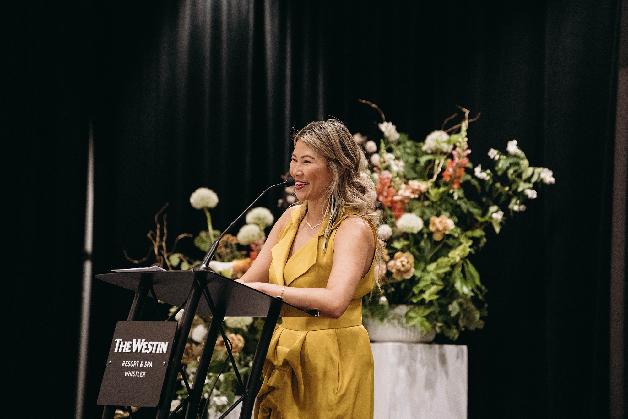 A woman in a sleeveless yellow dress stands behind a podium with a microphone, smiling while speaking at an event. There is a large floral arrangement behind her.