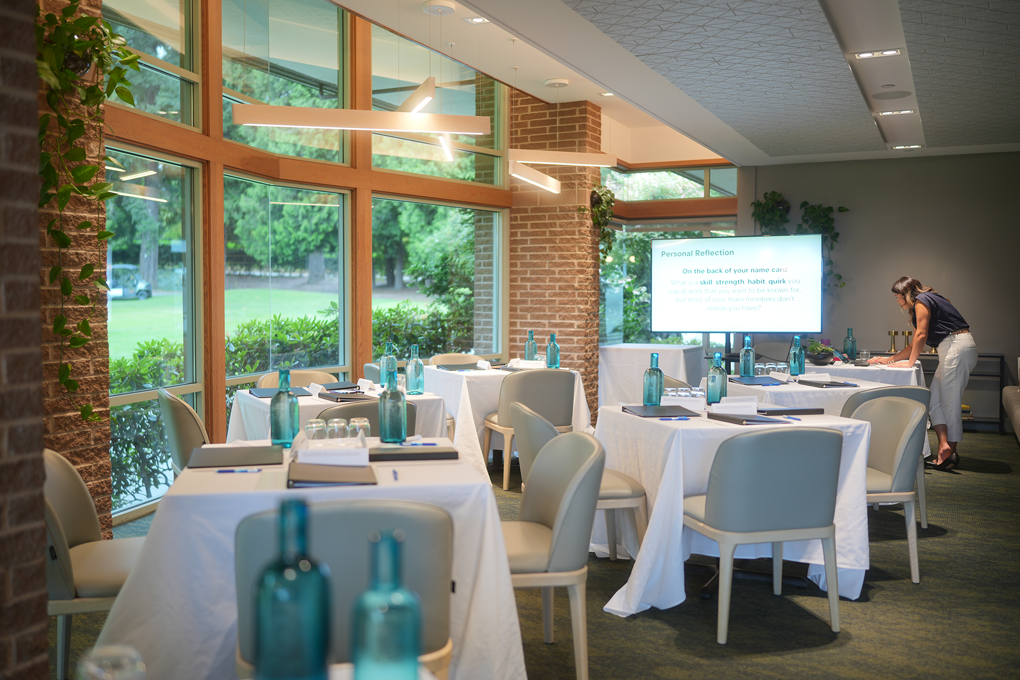 A modern conference or event room with large windows, brick and white walls, and a high ceiling with installed lights. The room is set up with white tablecloth-covered tables, each with blue glass bottles, glasses, notebooks, and writing materials. A woman is standing at one of the tables, arranging items. There is a large digital screen on the wall displaying a slide titled "Personal Reflection" with various questions for reflection.