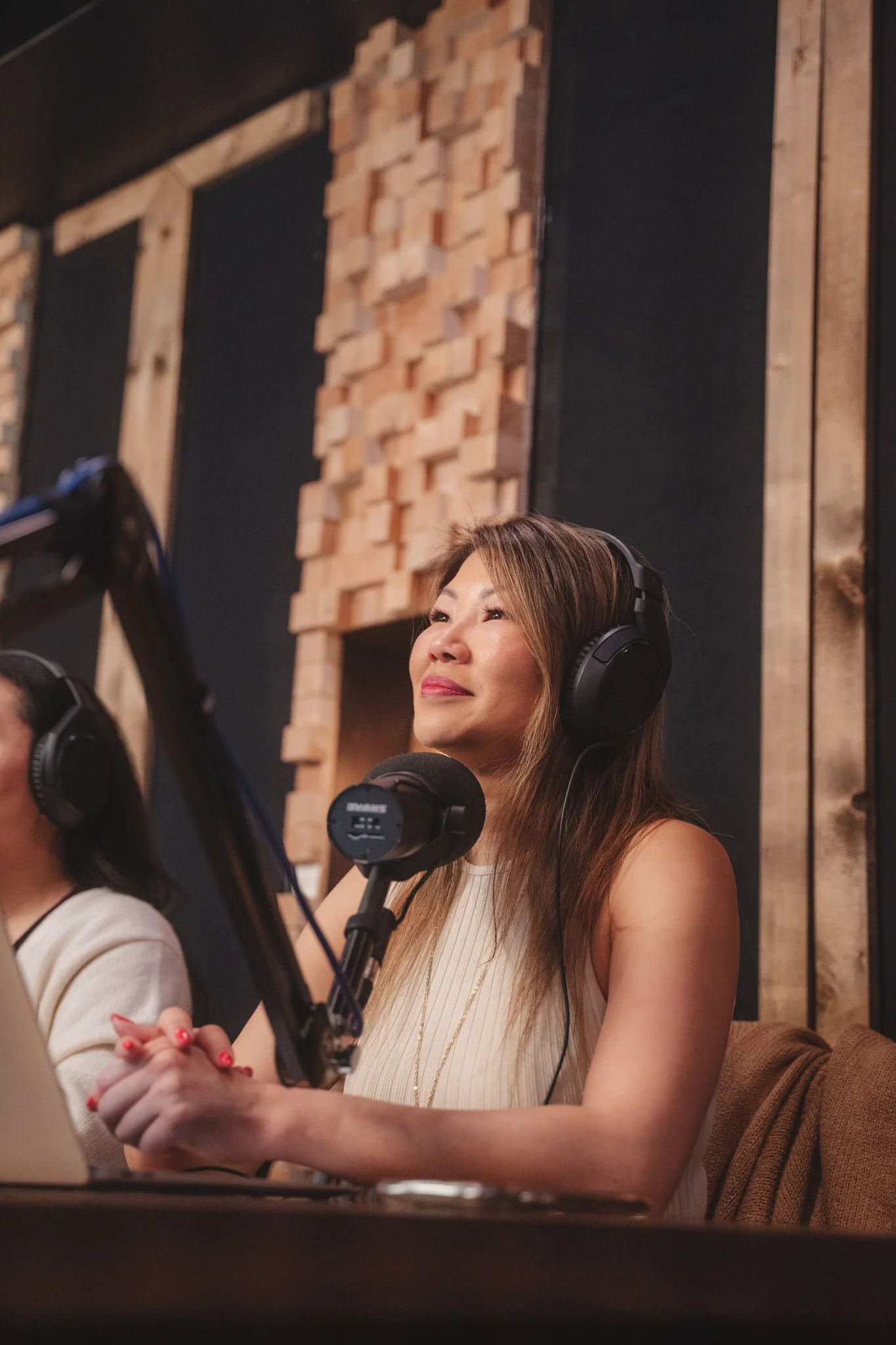 A woman with long brown hair wearing headphones and a cream-colored sleeveless top, sitting at a table in a recording studio, with her hands clasped together and looking thoughtfully. Acoustic wood paneling decorates the background.